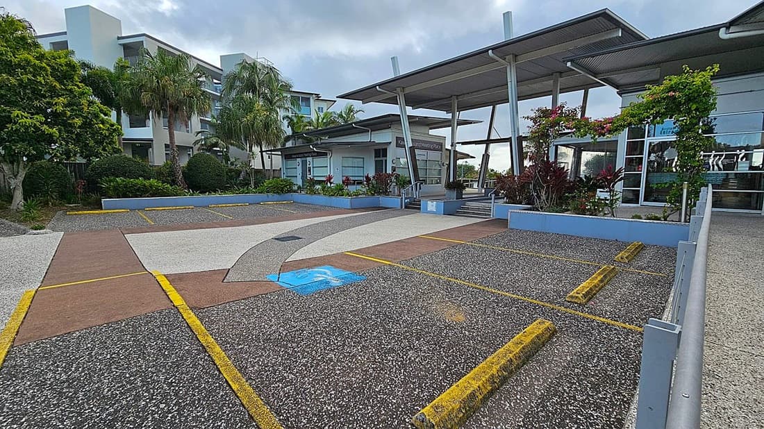 A modern building with a flat roof and large glass windows is on the right. In front, there are landscaped areas with shrubs and flowering plants. The foreground features a parking area with yellow lines and a designated blue disabled parking space. On the left, there are tall palm trees and a multi-storey building with balconies. The ground is a mix of textured concrete and asphalt.