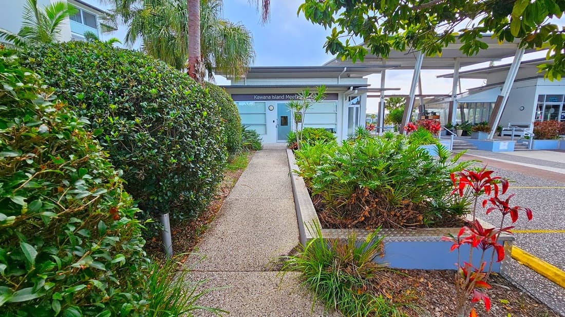 A pathway leads to a building with a sign reading "Kawana Island Meeting Place." On the left, dense green hedges and palm trees line the path. On the right, there are various plants, including ferns and red-leaved shrubs, in a raised garden bed. The building has large windows and a modern design. In the background, there are more structures with open seating areas and additional greenery. The ground is covered with a textured, speckled surface.