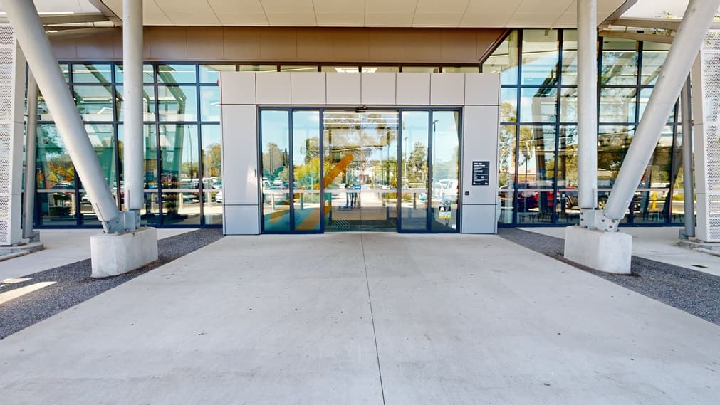 The entrance to the facility features double automatic glass sliding doors adorned with stickers. Glass walls surround the front and sides, while large concrete posts are visible to the left. In the foreground lies a spacious concrete concourse.