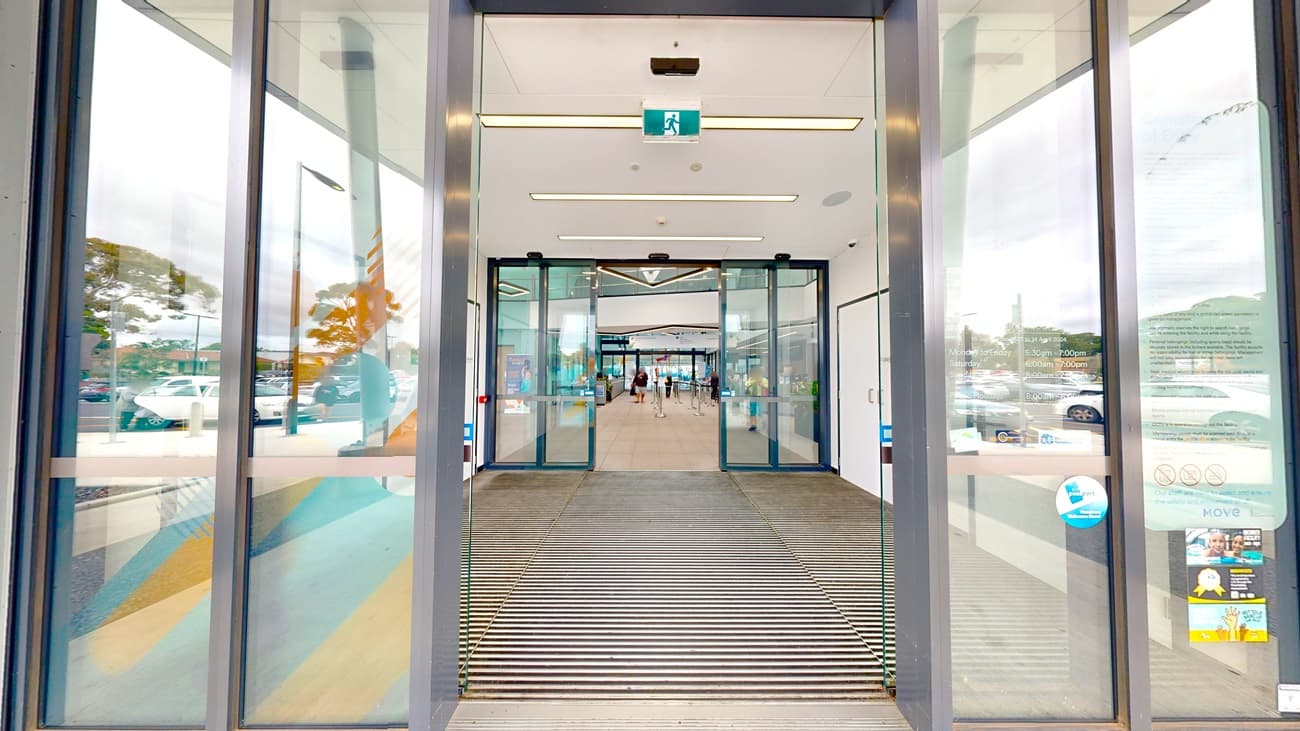 The facility entrance is outfitted with double automatic glass sliding doors, separated by a non-slippery mat. Stickers and posters are affixed to the glass wall. The second door leads into the building's interior. White tiles and a white ceiling with stylish light fixtures adorn the interior. Visitors are also present.