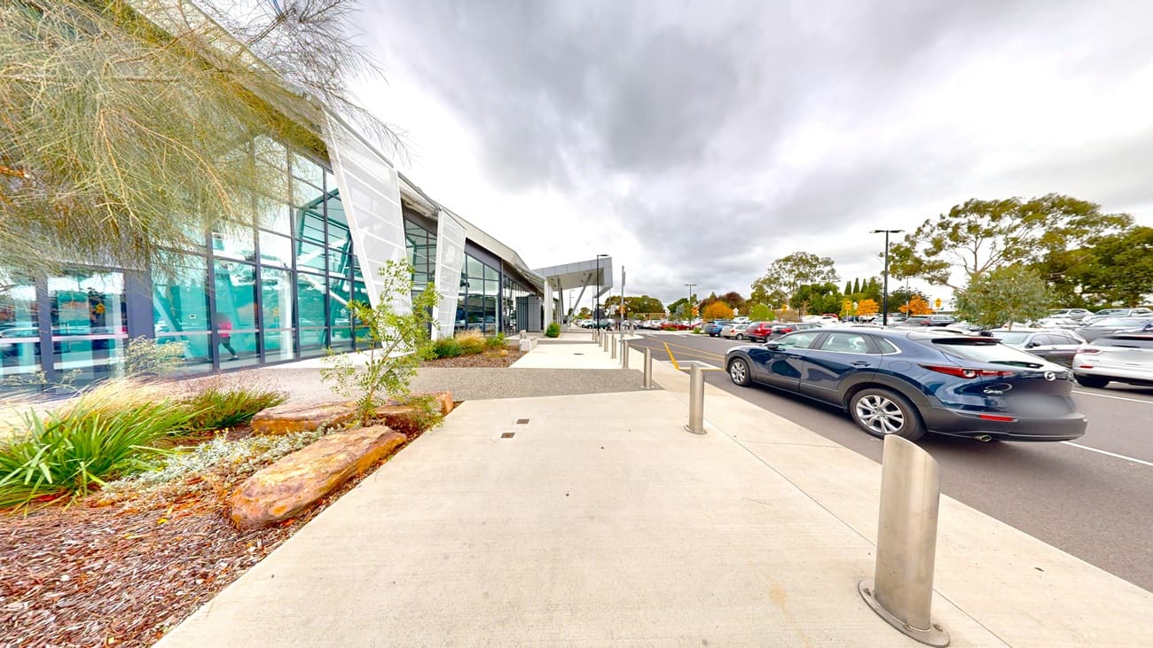 To the left stands a two-storey building with a glass facade and an extended front. Adjacent is a car park and driveway filled with various vehicles. Landscape plants, trees, and metal bollards line the building's front.