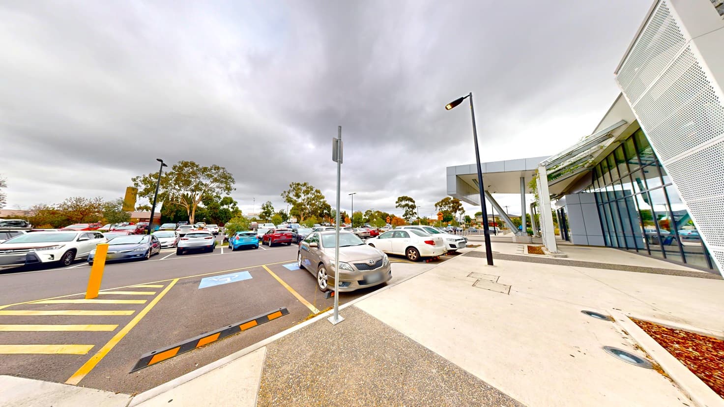 On the left, a car park is filled with various vehicles. To the right stands a two-story building with a glass facade and metal mesh accent, extending forward. In front is an accessible car park marked by signage. Streetlights and signs line the front, with tall trees visible in the background.