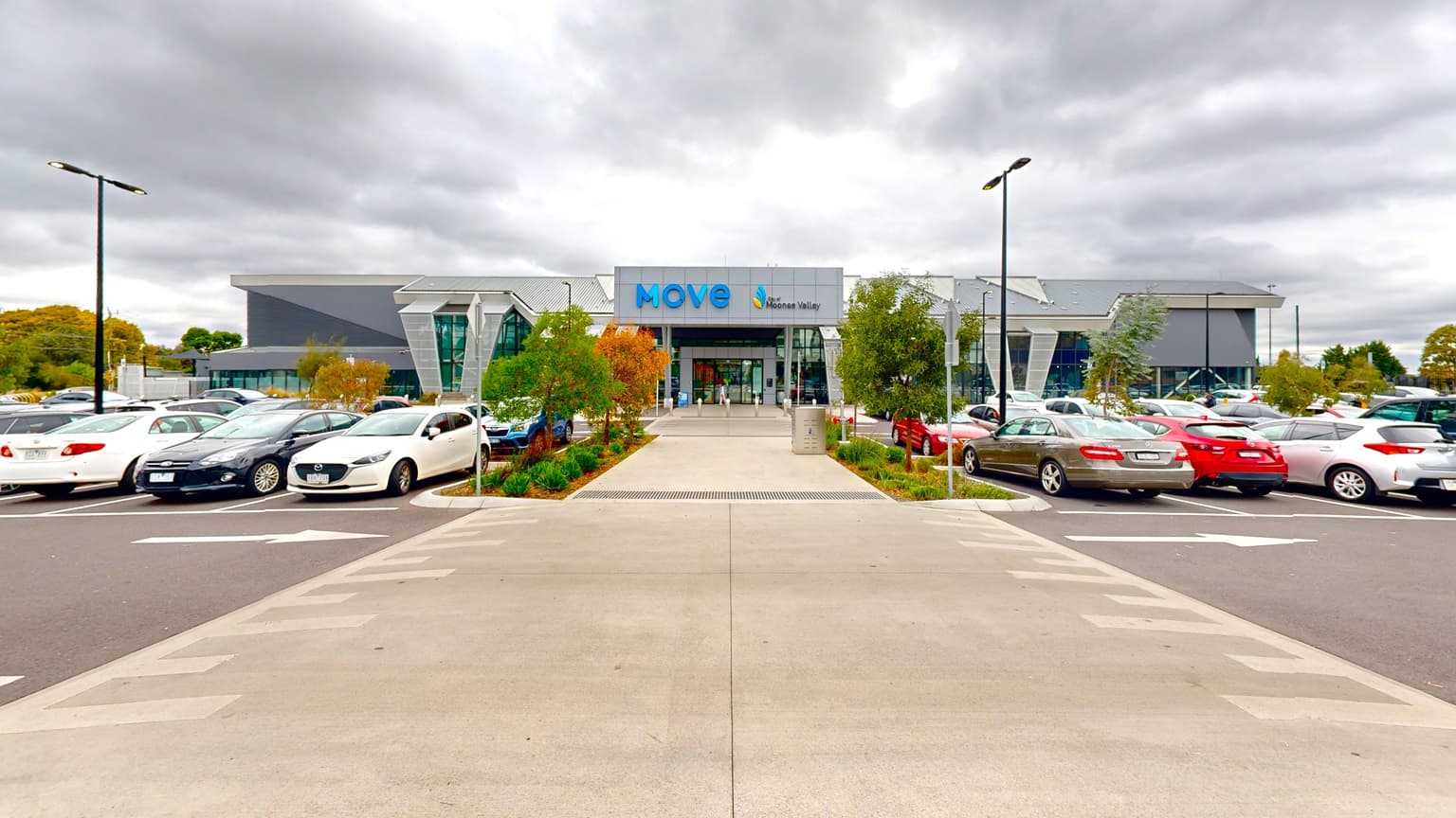 A modern building with a glass facade and signage reading "move" sits in the centre, flanked by parked cars on both the left and right. The left shows a line of cars in parking spaces, while the right has spaces with various makes of cars. A pedestrian crossing leads to the entrance, bordered by short green shrubbery and young trees. The car park extends to both sides, with the asphalt marked with white lines and parking bays.
