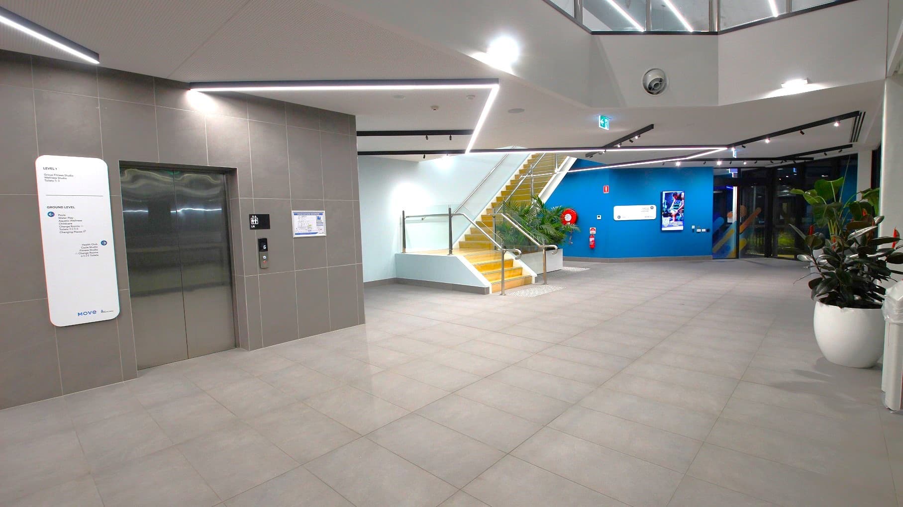 On the left, there is a lift with a grey tiled wall and a white information board beside it. In the middle, a staircase with yellow steps and glass railings leads upwards. On the right, a blue wall features a digital display and a fire hose reel. The floor is tiled in light grey, and there are large white planters with green plants. The ceiling has recessed lighting and a modern design with angular lines.