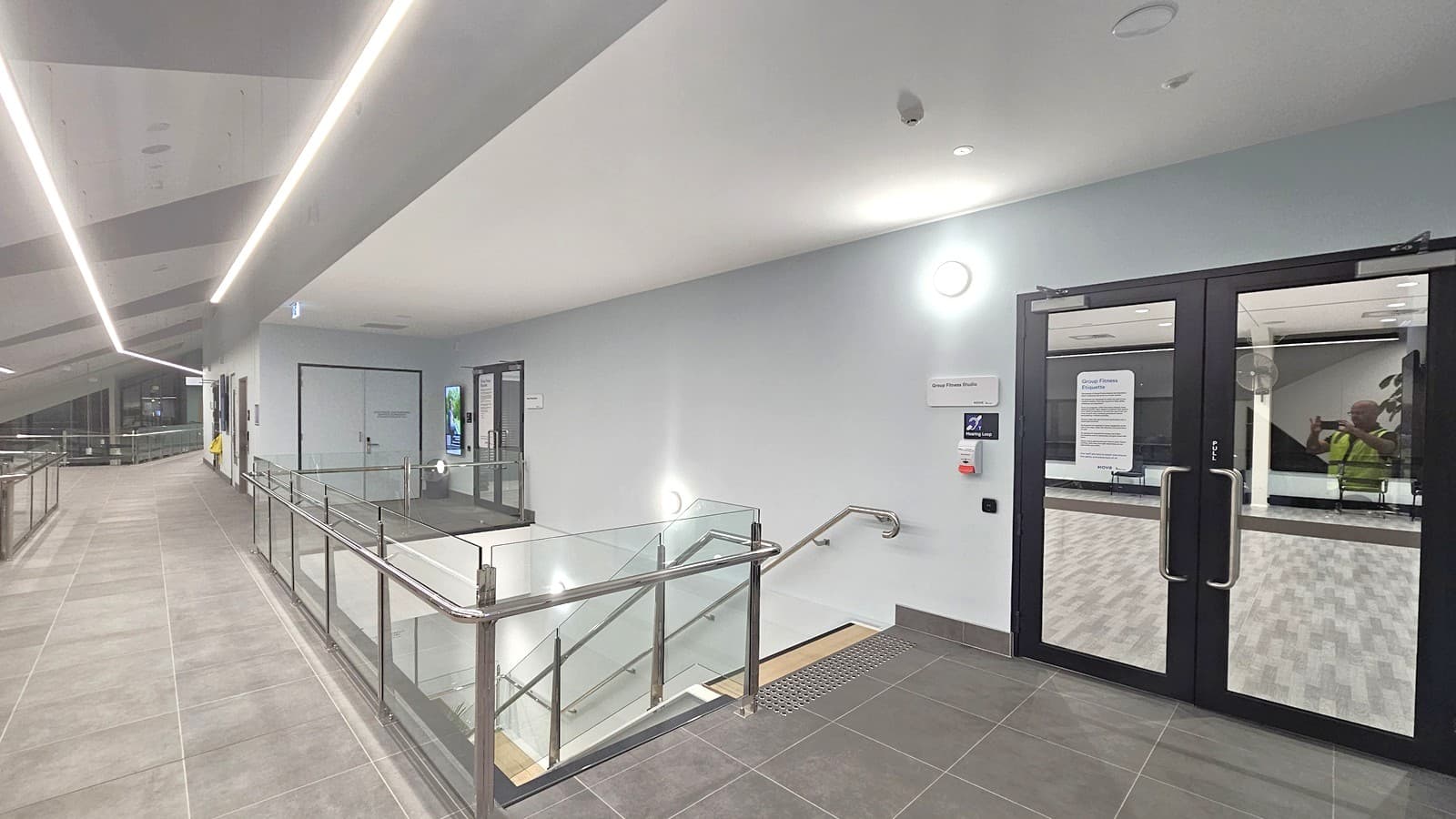 A modern indoor corridor with a tiled floor and a ceiling featuring linear lights. On the left, a walkway with a glass railing leads to a distant area. On the right, there are double glass doors with notices attached, and a staircase with a metal handrail descends. The walls are plain, with a few wall-mounted lights. A sign and a small digital screen are visible near the doors.