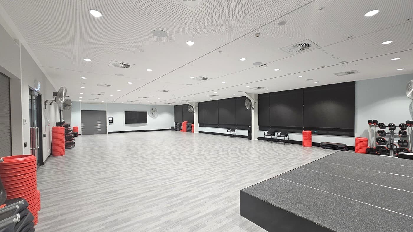 A spacious gym room with a light wooden floor and a white ceiling with recessed lighting. On the left, there are stacks of red exercise steps and a wall-mounted fan. The back wall features large black panels and more red exercise steps. On the right, there are racks of dumbbells and additional red steps. A raised platform is in the foreground on the right side. The room is well-lit and designed for fitness activities.
