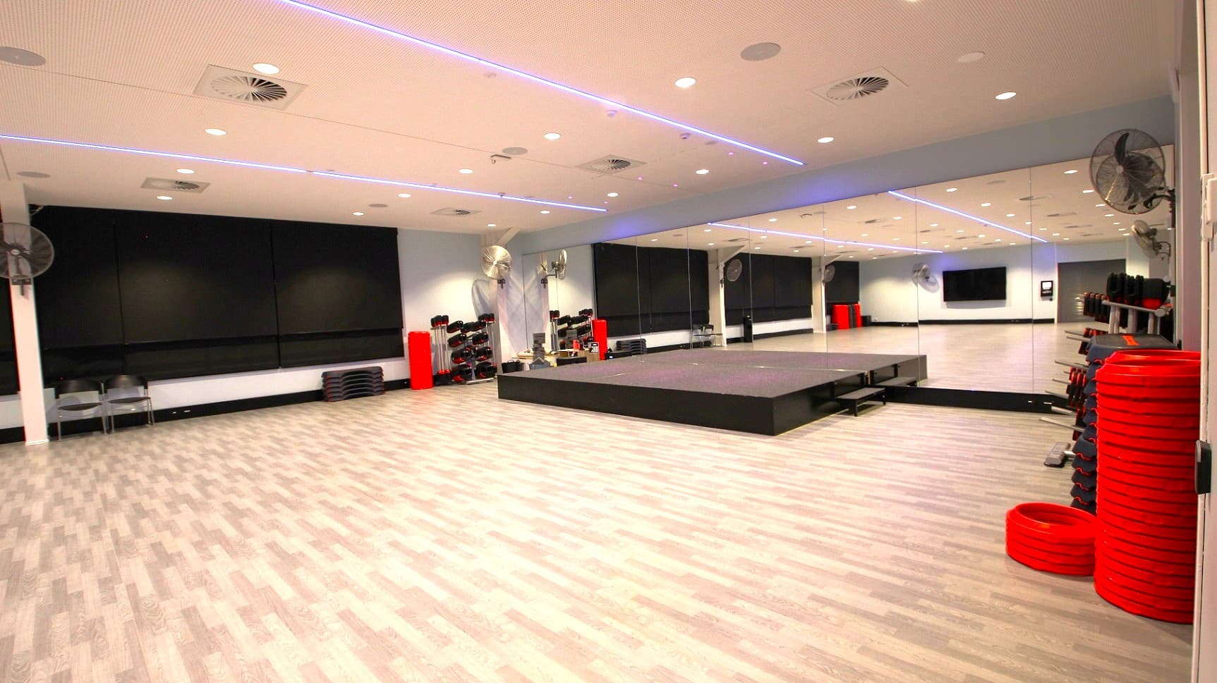 A spacious gym studio with a light wood-patterned floor. On the right, there are stacks of red weight plates and a mirrored wall reflecting the room. In the centre, a raised platform is visible. The left side has black mats and chairs against the wall. The ceiling is white with recessed lighting and blue LED strips. Fans are mounted on the walls.