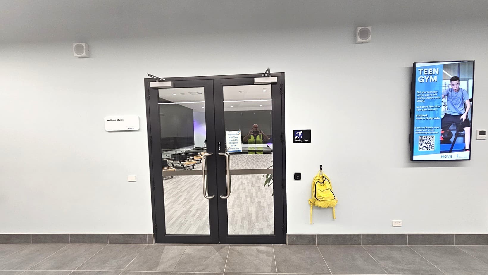 A pair of glass doors in the centre with metal handles, leading to a room with light wood flooring. To the left of the doors, a sign reads "Wellness Studio". On the right, a digital screen displays an advertisement for a "Teen Gym" with a QR code. Below the screen, a bright yellow backpack hangs on a hook. The floor is tiled in grey, and the walls are light-coloured. Ceiling speakers are visible above.