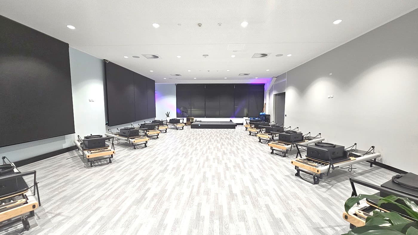 A spacious room with a light grey wooden floor and white ceiling with recessed lighting. On the left and right, there are rows of black exercise reformers with wooden accents. The back wall features large black panels, and a small stage is centred against it. A plant is visible in the foreground on the right side. The room has a modern and minimalist design.