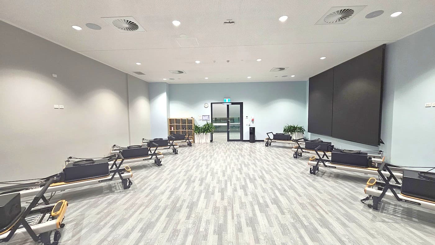 A spacious room with a light grey wooden floor and a white ceiling featuring recessed lighting and air vents. On the left and right sides, there are several black exercise reformer machines lined up. The back wall has a large black screen on the right and a glass door in the centre, flanked by potted plants and a small shelving unit on the left. A clock and a small wall-mounted device are visible near the door.