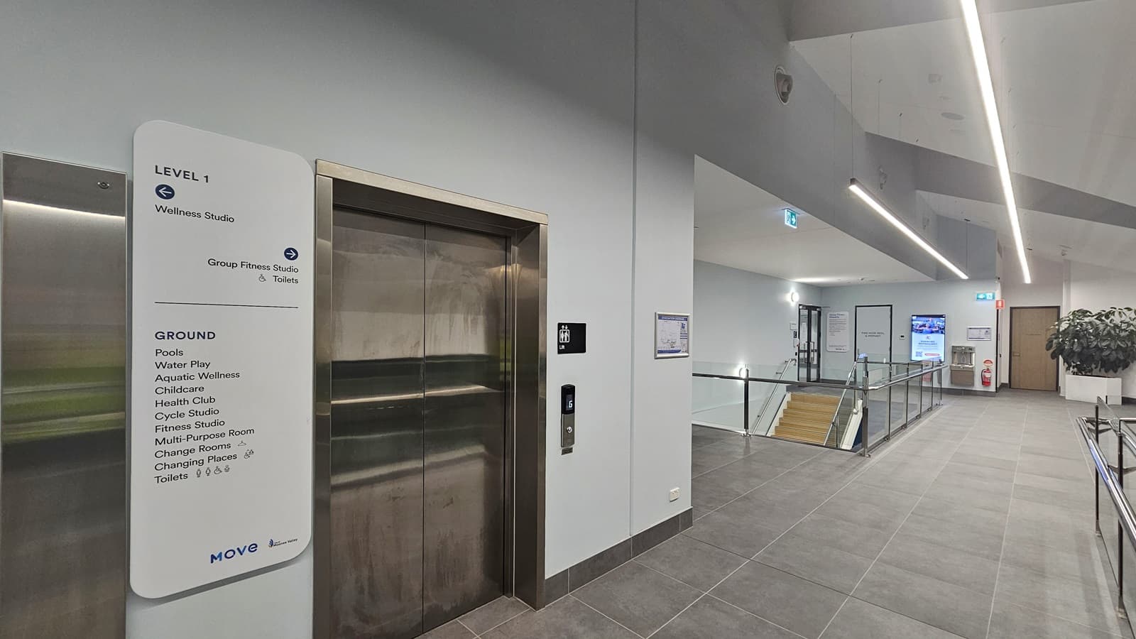 On the left, there is a lift with a metallic door and a sign listing facilities on different levels, including a wellness studio and fitness areas. In the middle, a corridor leads to a staircase with a glass railing. On the right, there is a digital display, a fire extinguisher, and a potted plant. The floor is tiled in grey, and the ceiling features linear lights.
