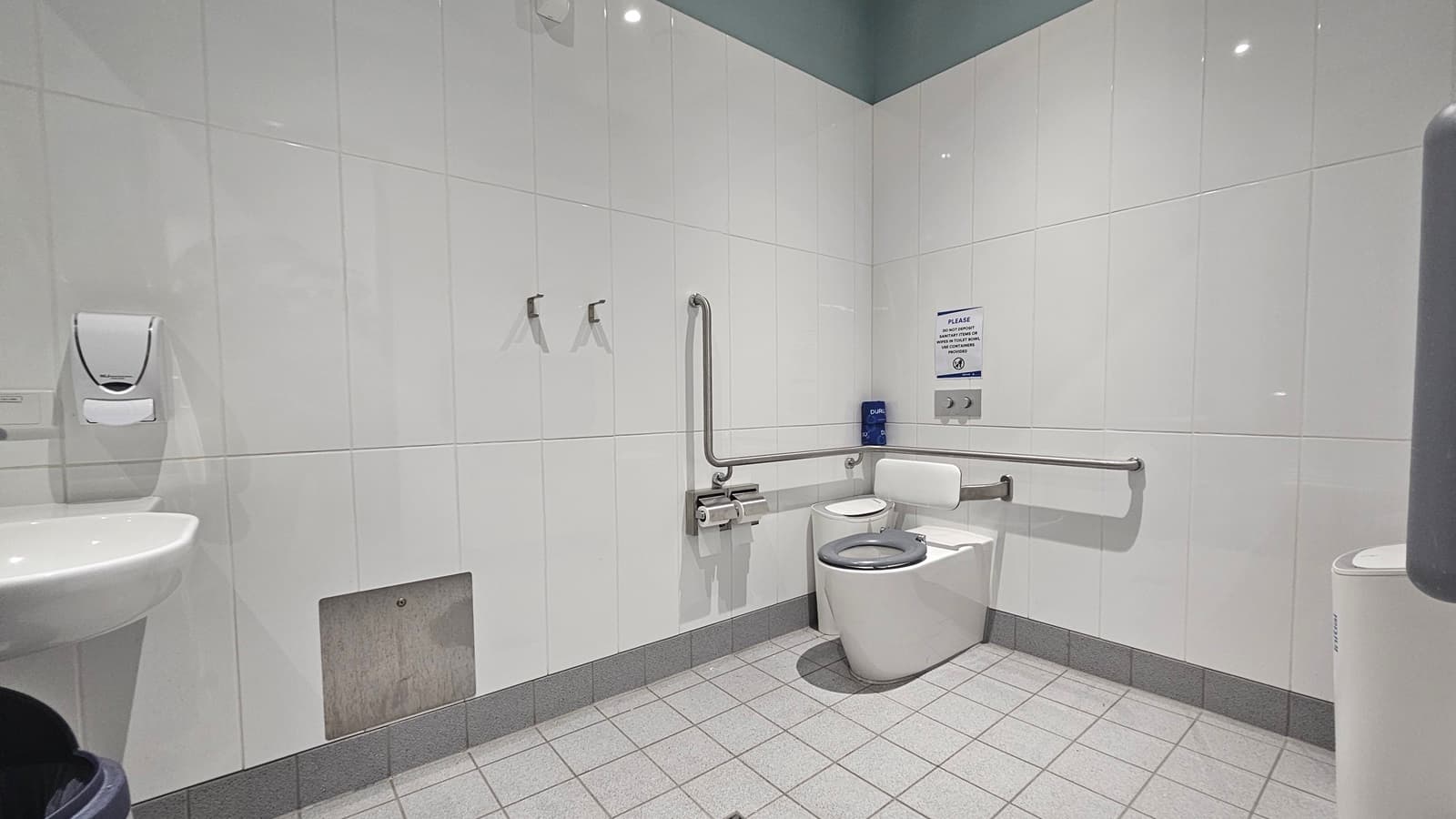 A public restroom with white tiled walls and a light grey tiled floor. On the left, there's a white sink with a soap dispenser above it. In the centre, two metal hooks are mounted on the wall. On the right, a toilet with a backrest is equipped with grab bars for accessibility. Above the toilet, a sign and a blue dispenser are visible. The ceiling is light green, and a waste bin is partially visible in the foreground.