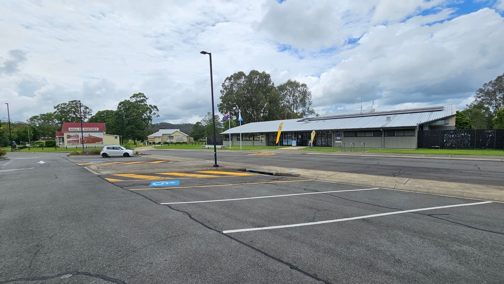 A curved road leads to a parking area with a few cars. On the left, a single-storey building with a metal roof and several flags in front is surrounded by grass. The building has large windows and a modern design. In the centre, a streetlamp stands on a small landscaped island with plants. The right side features more parking spaces and trees in the background. The road surface is cracked, and the area is open and spacious.