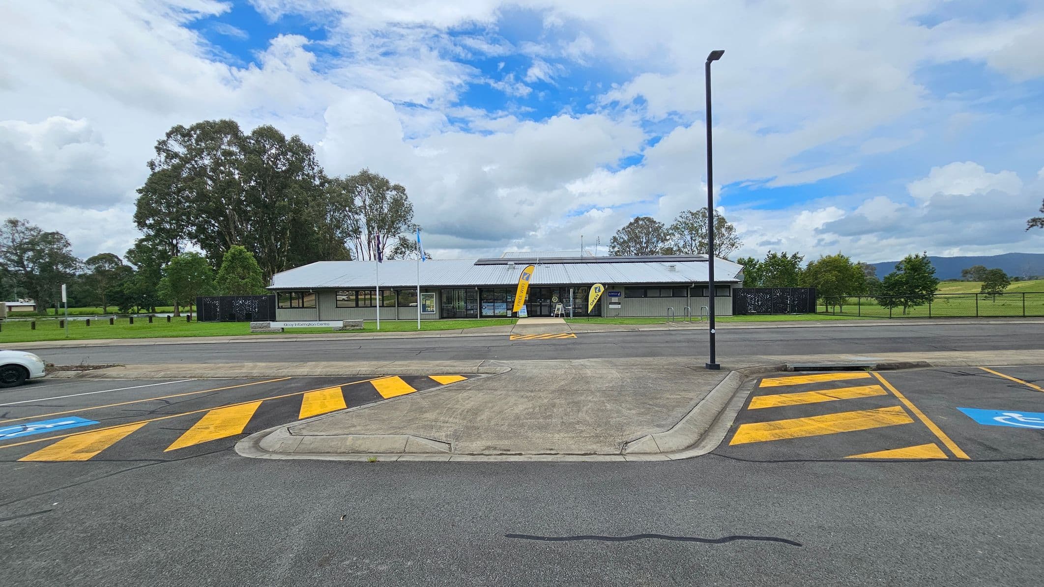 A single-storey building with a grey roof and large windows is in the centre, with two yellow flags at the entrance. In the foreground, there is a car park with yellow and black striped markings and a blue disabled parking sign. A white car is partially visible on the left. The background features a grassy area with tall trees and a mountain range in the distance. A tall streetlight stands on the right side of the image.