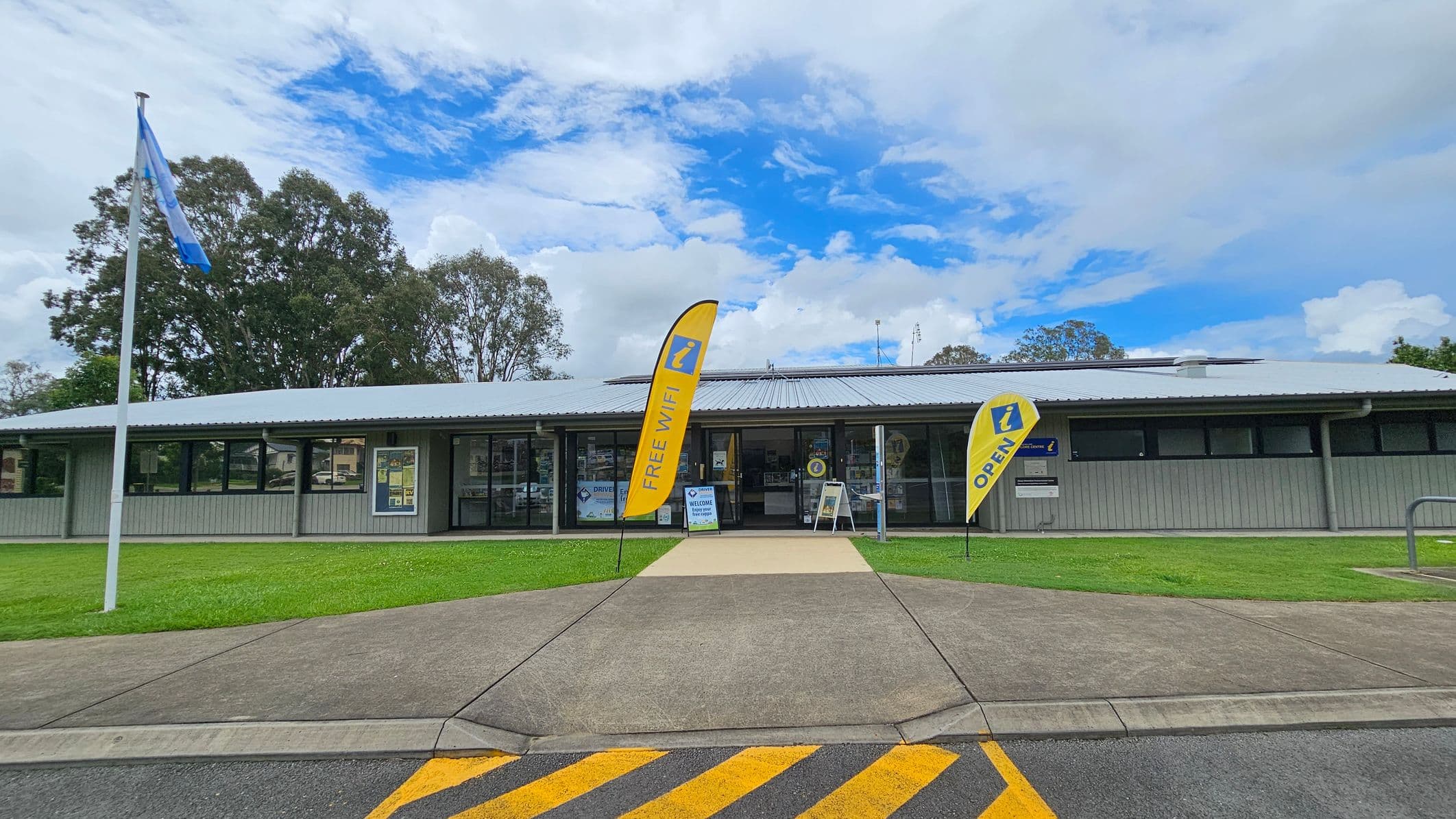 A single-storey building with a grey metal roof and wooden exterior. In front, two yellow flags display "FREE WIFI" and "OPEN". A concrete path leads to the entrance, flanked by grass. A flagpole with a blue flag stands on the left. The foreground features a road with yellow diagonal markings. Trees are visible in the background.