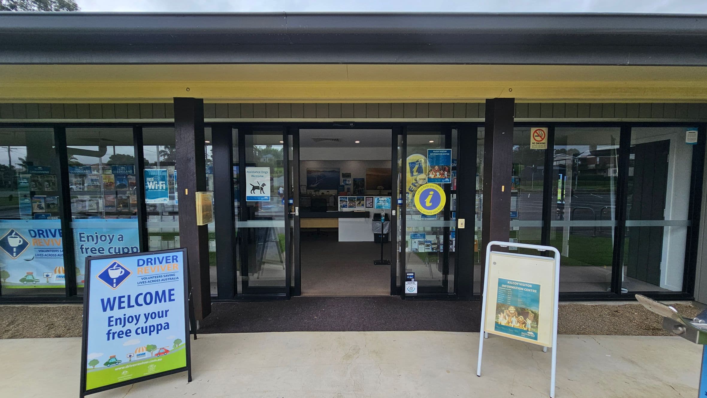 Entrance to a visitor information centre with glass doors. A sign on the left reads "WELCOME Enjoy your free cuppa" with a "Driver Reviver" logo. Another sign on the right displays information about the centre. Inside, brochures and posters are visible on the walls. The floor is concrete outside and carpeted inside. The ceiling is plain with lighting fixtures. A water fountain is partially visible on the right.