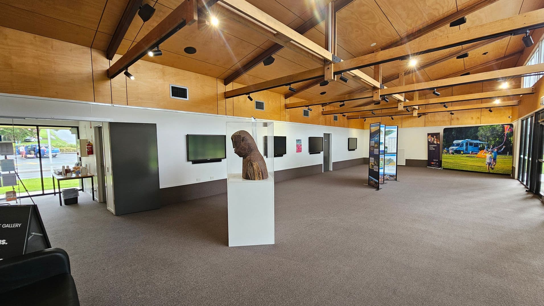 A spacious gallery with a high wooden ceiling featuring exposed beams and spotlights. On the left, a glass door reveals an outdoor view with a parked car. Inside, a table with brochures is near the entrance. In the centre, a wooden sculpture is displayed on a white pedestal. The right side has a large photo of a family camping scene. Several screens and informational panels are mounted on the walls. The floor is carpeted in a neutral tone.