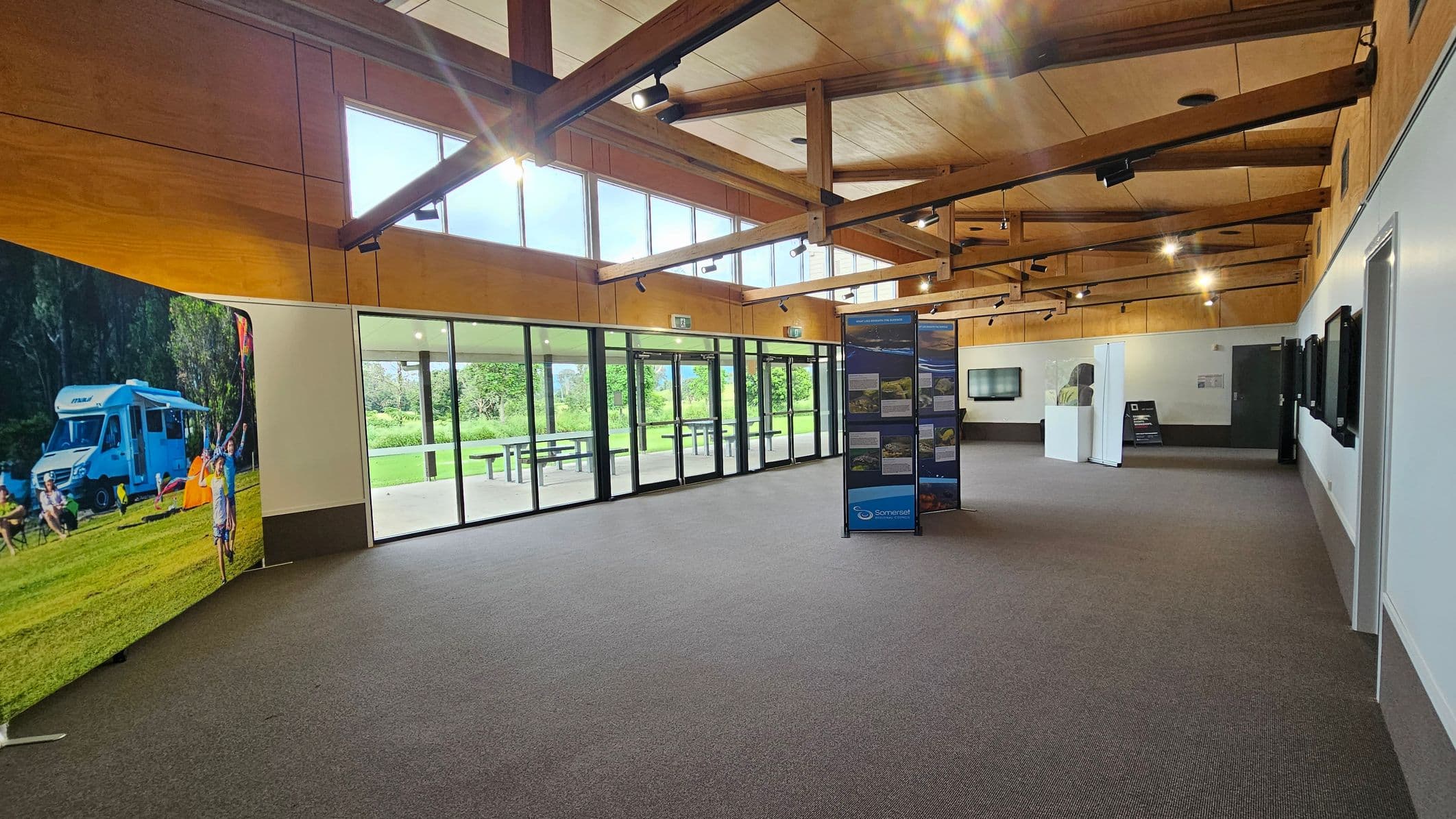 Interior of a spacious room with a grey carpeted floor and a wooden ceiling featuring exposed beams. On the left, a large display shows an outdoor scene with a camper van and people. The middle has a series of glass doors and windows, offering a view of greenery and picnic tables outside. In the centre, a stand with informational panels is visible. The right side has a white wall with mounted screens and a doorway. The room is well-lit with ceiling lights.