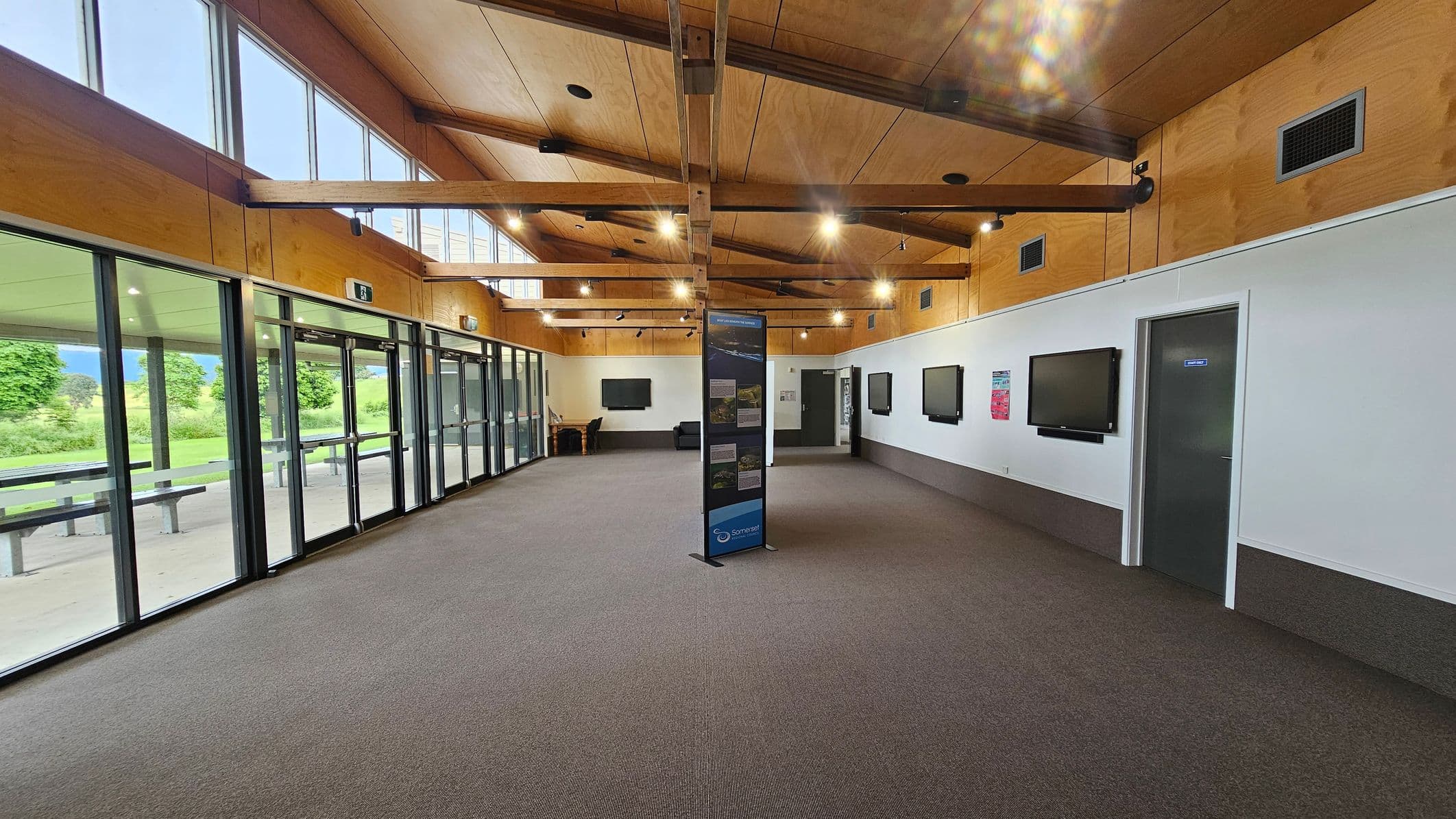 A spacious room with a high wooden ceiling and exposed beams. On the left, large glass doors and windows offer a view of a green outdoor area with trees and picnic tables. The right wall is white with several mounted screens and a grey door. In the centre, a freestanding display board is present. The floor is carpeted in a mix of grey and brown tones. Ceiling lights are evenly spaced, providing ample illumination.