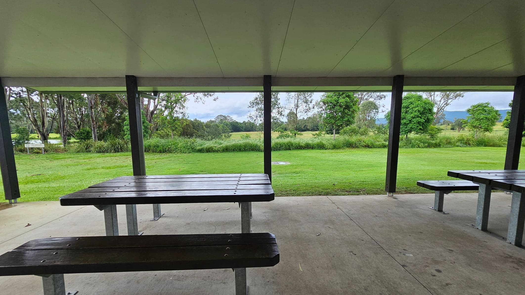A sheltered picnic area with wooden tables and benches on a concrete floor. The ceiling is flat and light-coloured. In the background, there is a grassy field with several trees and shrubs. Beyond the field, more trees and a distant view of hills or mountains are visible. The area is open and surrounded by greenery.