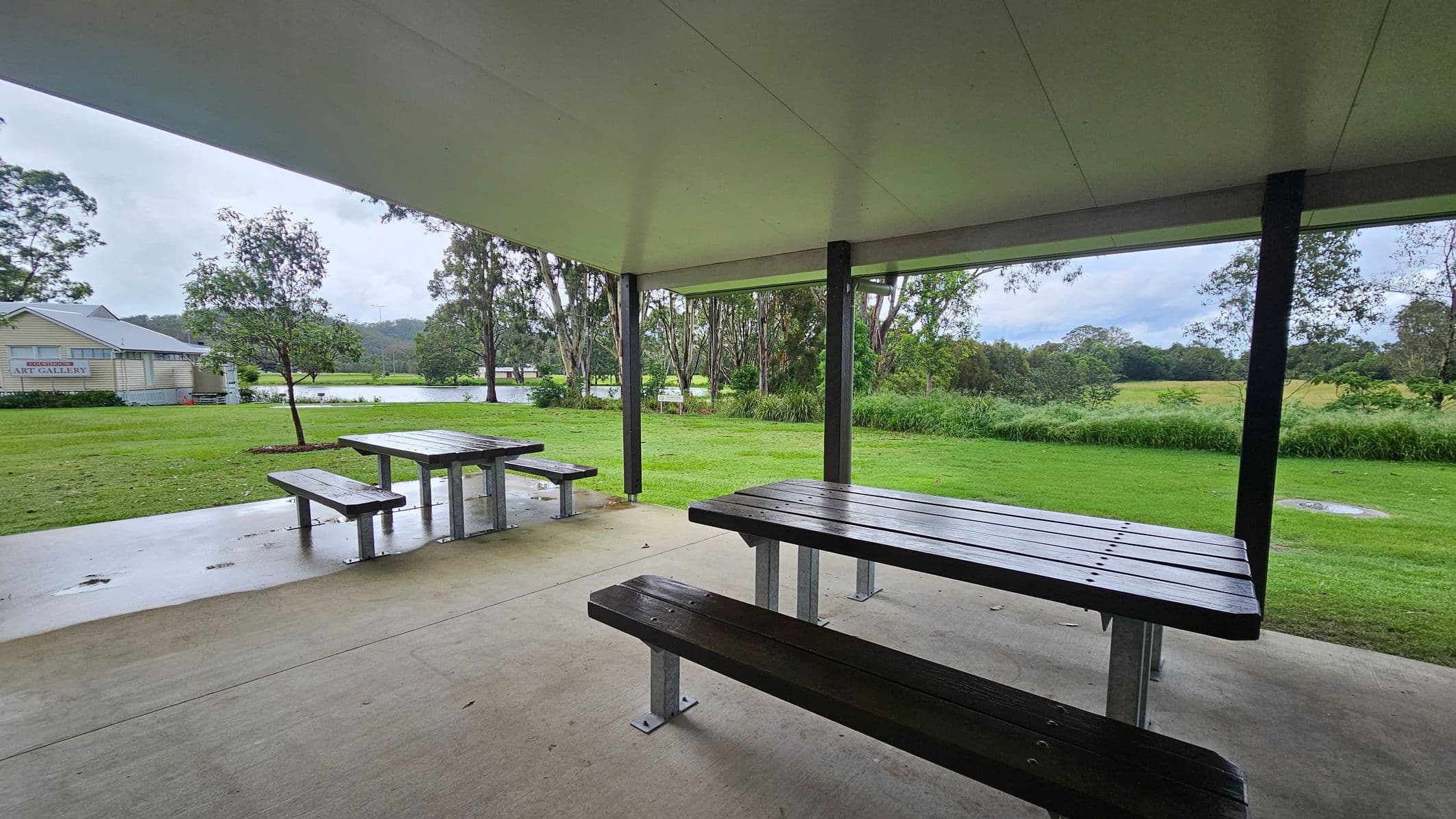 Under a shelter with a white ceiling, there are two wooden picnic tables on a concrete floor. The foreground shows a table with benches, while another is visible to the left. Beyond the shelter, a grassy area extends to a small art gallery building on the left. Trees and a body of water are visible in the background, with more greenery and hills in the distance.