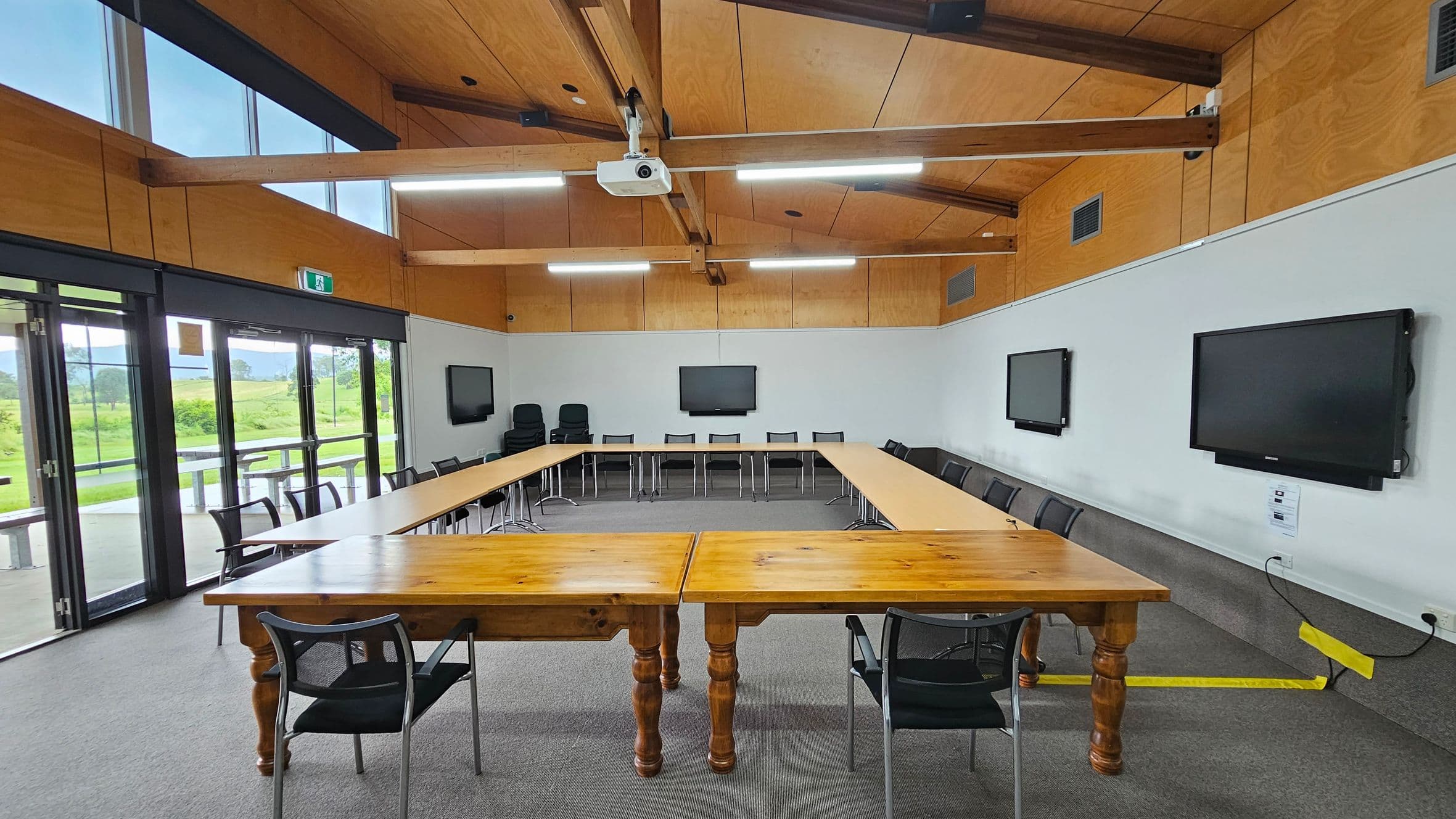 A conference room with a U-shaped arrangement of wooden tables and black chairs. On the left, there's a whiteboard on a stand. The back wall features large windows and glass doors leading outside, allowing natural light to enter. The right wall has two mounted screens. The ceiling is wooden with exposed beams and fluorescent lights. The floor is carpeted in grey.