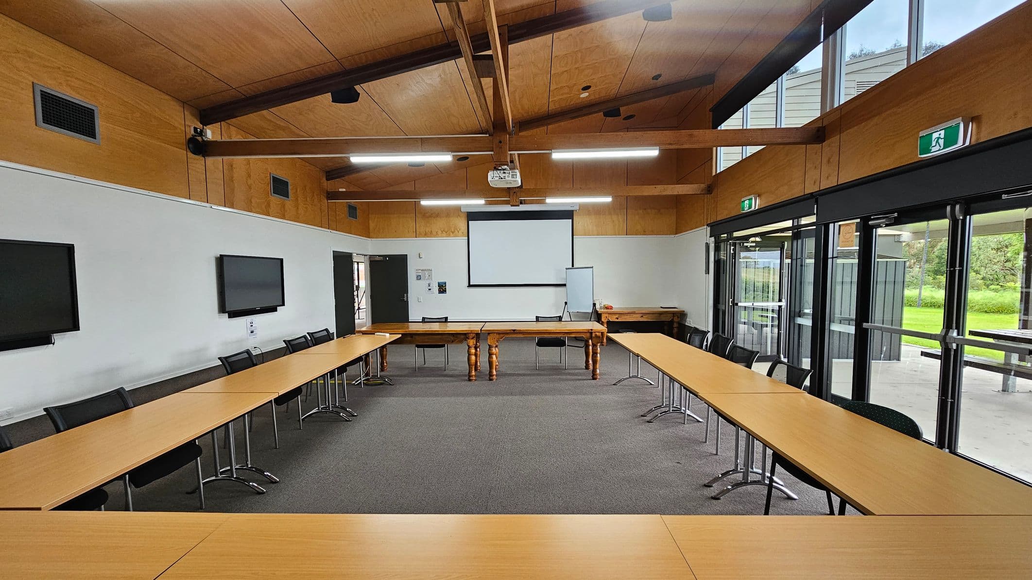 A conference room with a U-shaped arrangement of wooden tables and black chairs. The back wall features a large projection screen and a whiteboard. Two flat-screen monitors are mounted on the left wall. The right side has large glass doors with views of greenery outside. The ceiling is wooden with exposed beams and fluorescent lights. The floor is carpeted in grey.