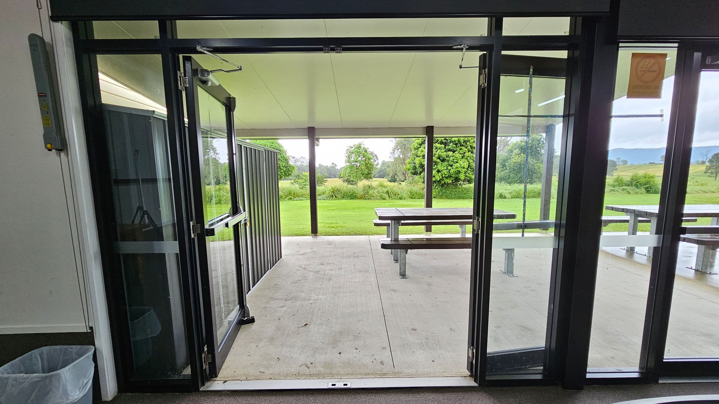 Glass doors open to a covered outdoor area with a concrete floor. In the middle, there are picnic tables with benches. Beyond the covered area, a grassy field extends into the distance, bordered by trees and shrubs. The ceiling of the covered area is plain and light-coloured. On the left, a waste bin is visible inside the building. The right side of the image shows a glass panel with a small sign.
