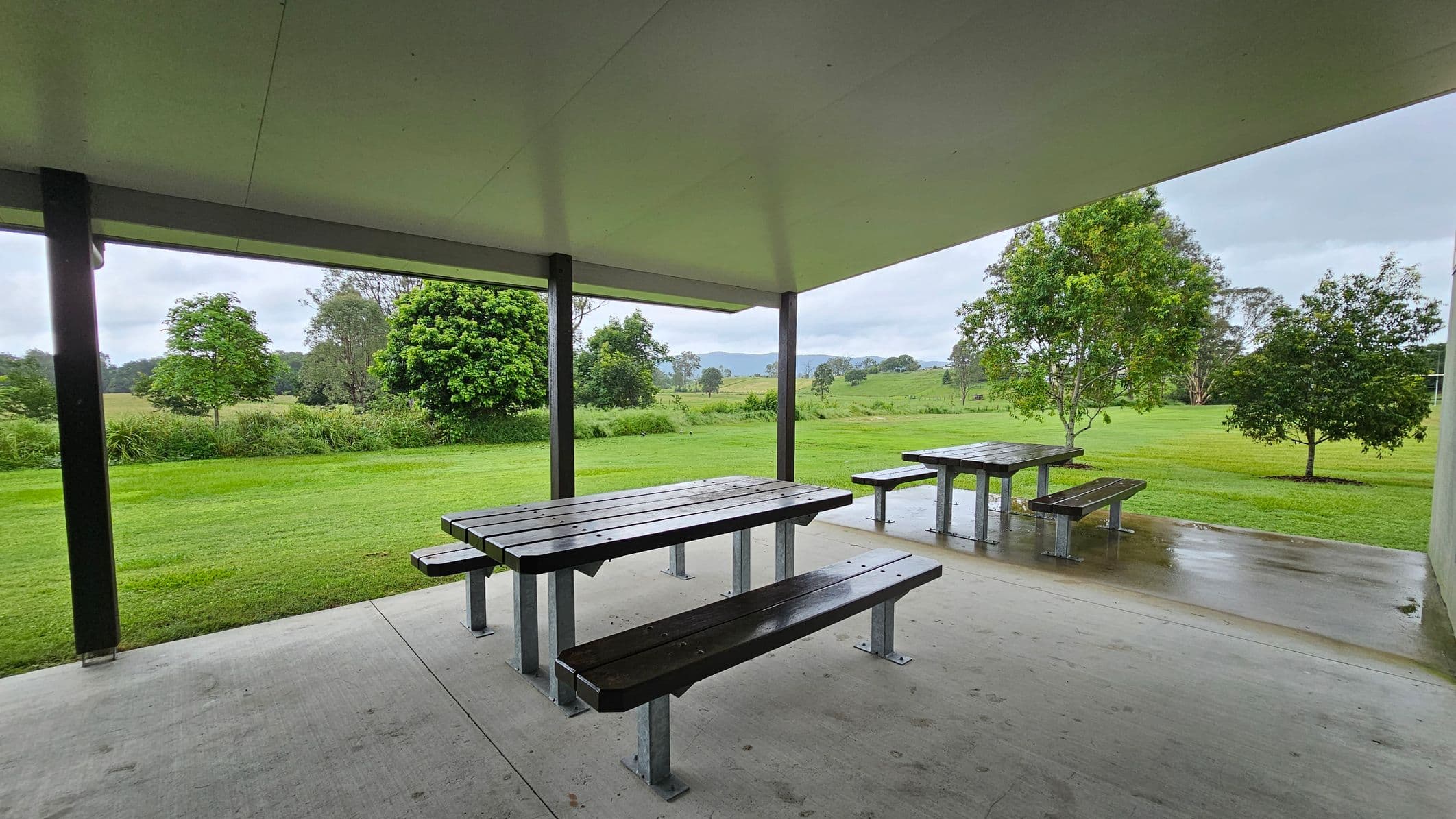 Under a shelter with a light-coloured ceiling, there are two picnic tables with benches on a concrete floor. The foreground shows a wet surface, suggesting recent rain. In the background, a lush green landscape with several trees and a grassy field extends into the distance. The scene is open and spacious, with a peaceful, natural setting.