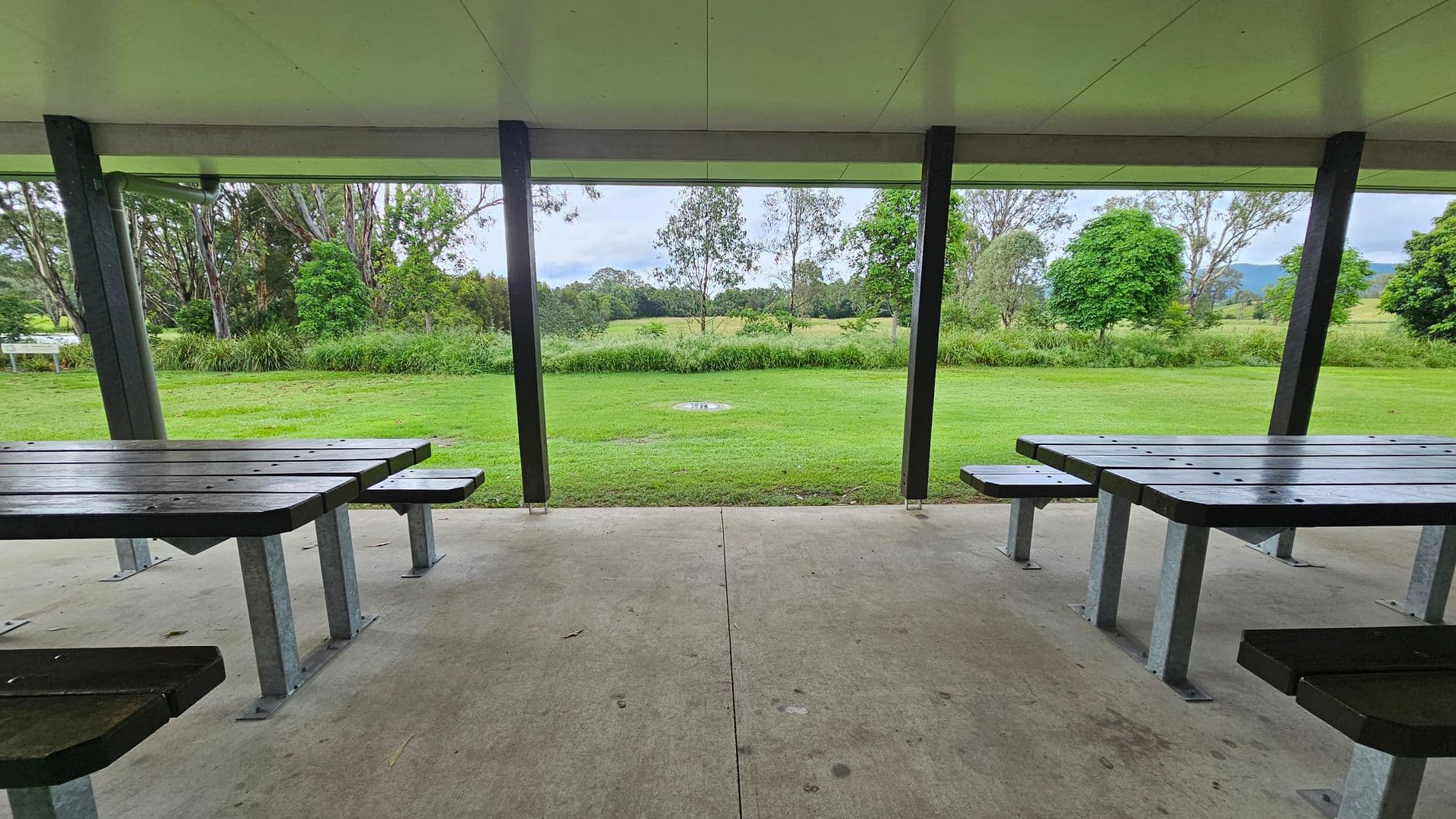Under a sheltered area with a flat ceiling, there are two picnic tables with attached benches on either side. The floor is concrete. Beyond the shelter, there's a grassy field with scattered trees and shrubs. In the distance, more trees and a hint of hills or mountains are visible. The scene is framed by vertical support beams of the shelter.