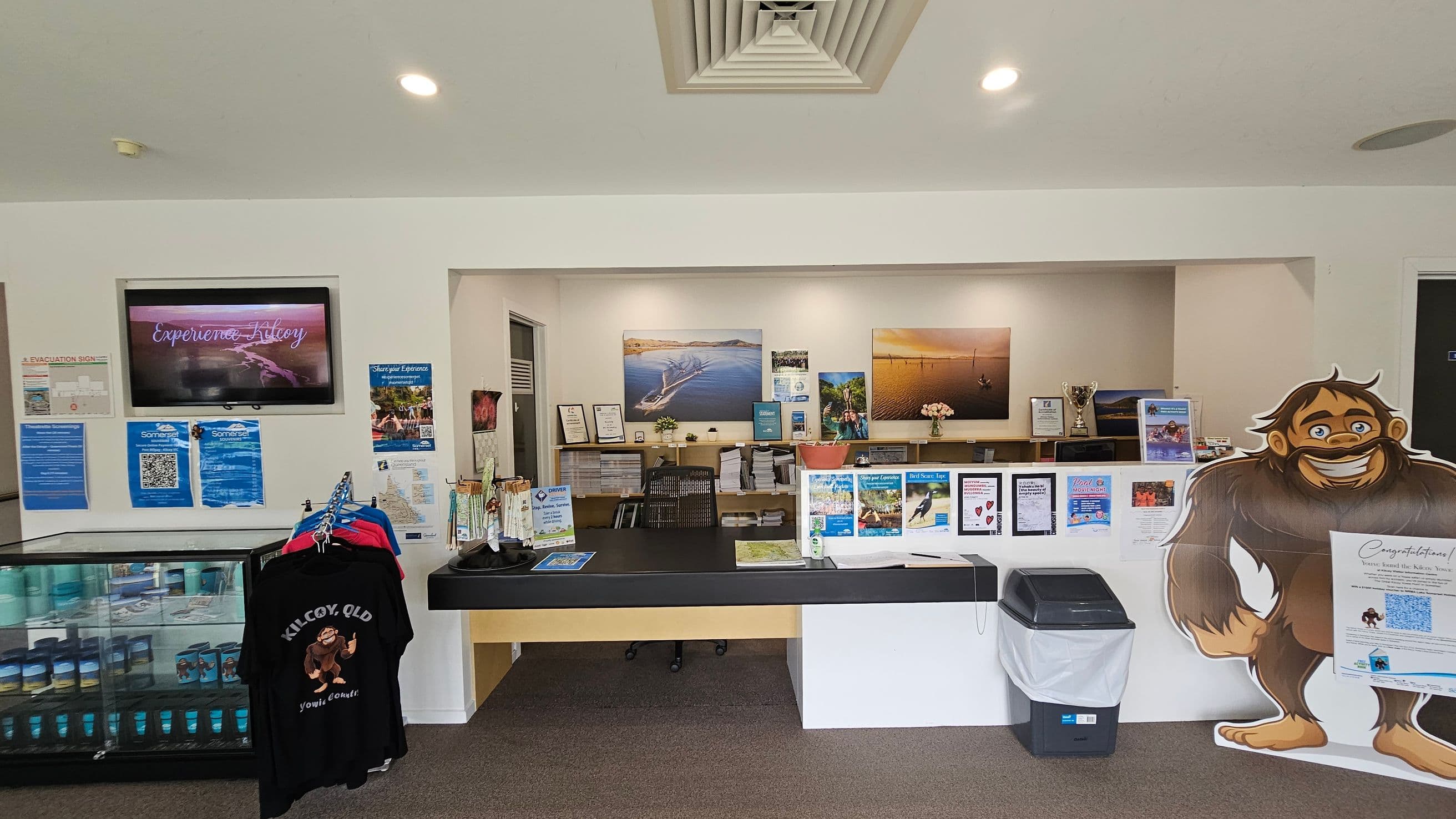 A reception area with a counter in the centre, displaying brochures and a map. Behind the counter are shelves with books, trophies, and framed pictures of landscapes. On the left, a wall-mounted screen shows "Experience Kilcoy" above a display of posters and a rack of black T-shirts. On the right, a large cut-out of a cartoon character stands next to a bin. The ceiling has recessed lighting and an air vent, and the floor is carpeted.