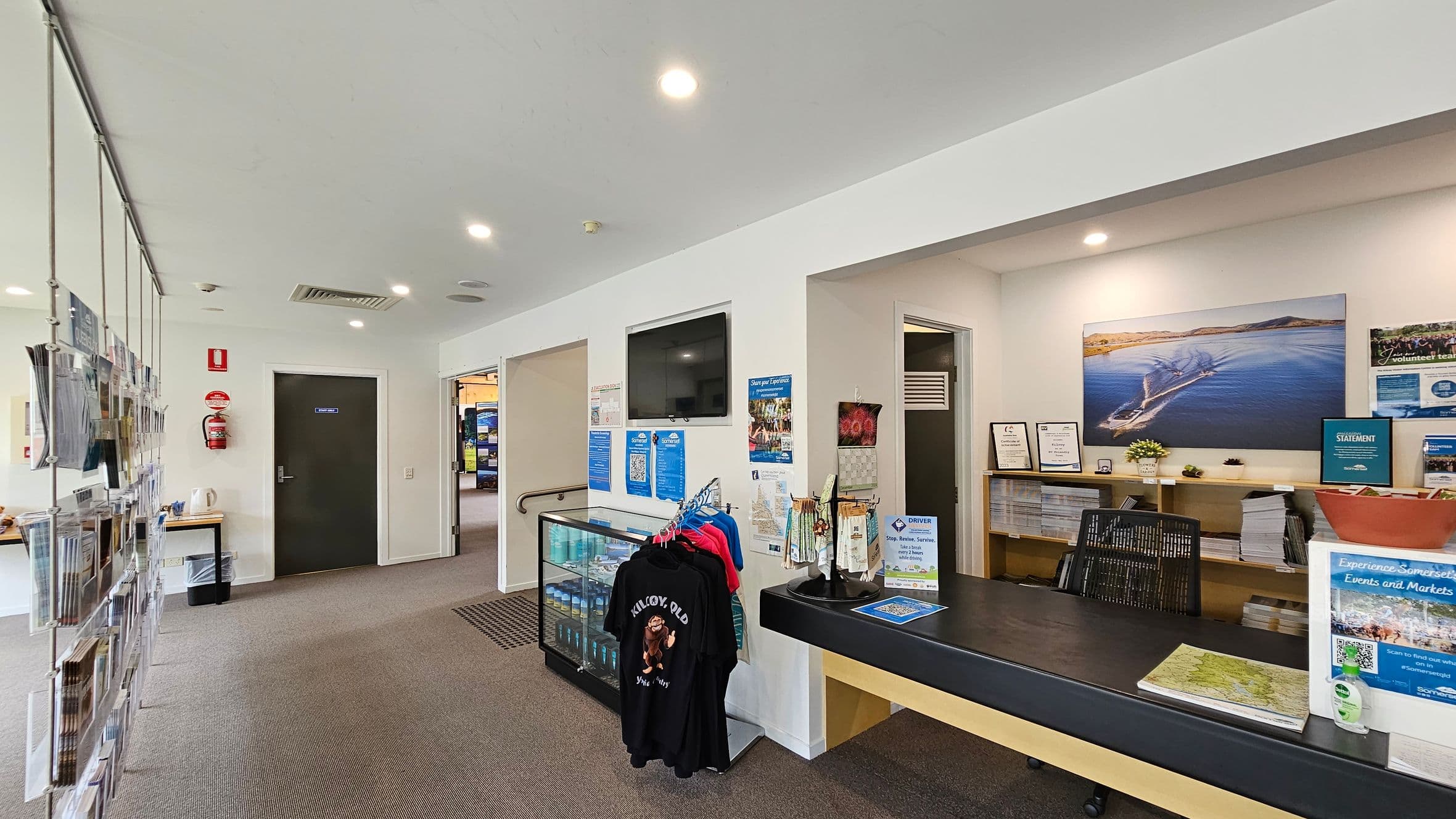 A reception area with a counter on the right displaying brochures, a map, and a hand sanitiser bottle. Behind the counter, shelves hold more brochures and small potted plants. A large photograph of a boat on water is mounted on the wall. In the centre, a rack with colourful T-shirts is visible. On the left, a wall-mounted display holds various pamphlets. The floor is carpeted, and the ceiling has recessed lighting. A door and a fire extinguisher are also visible.