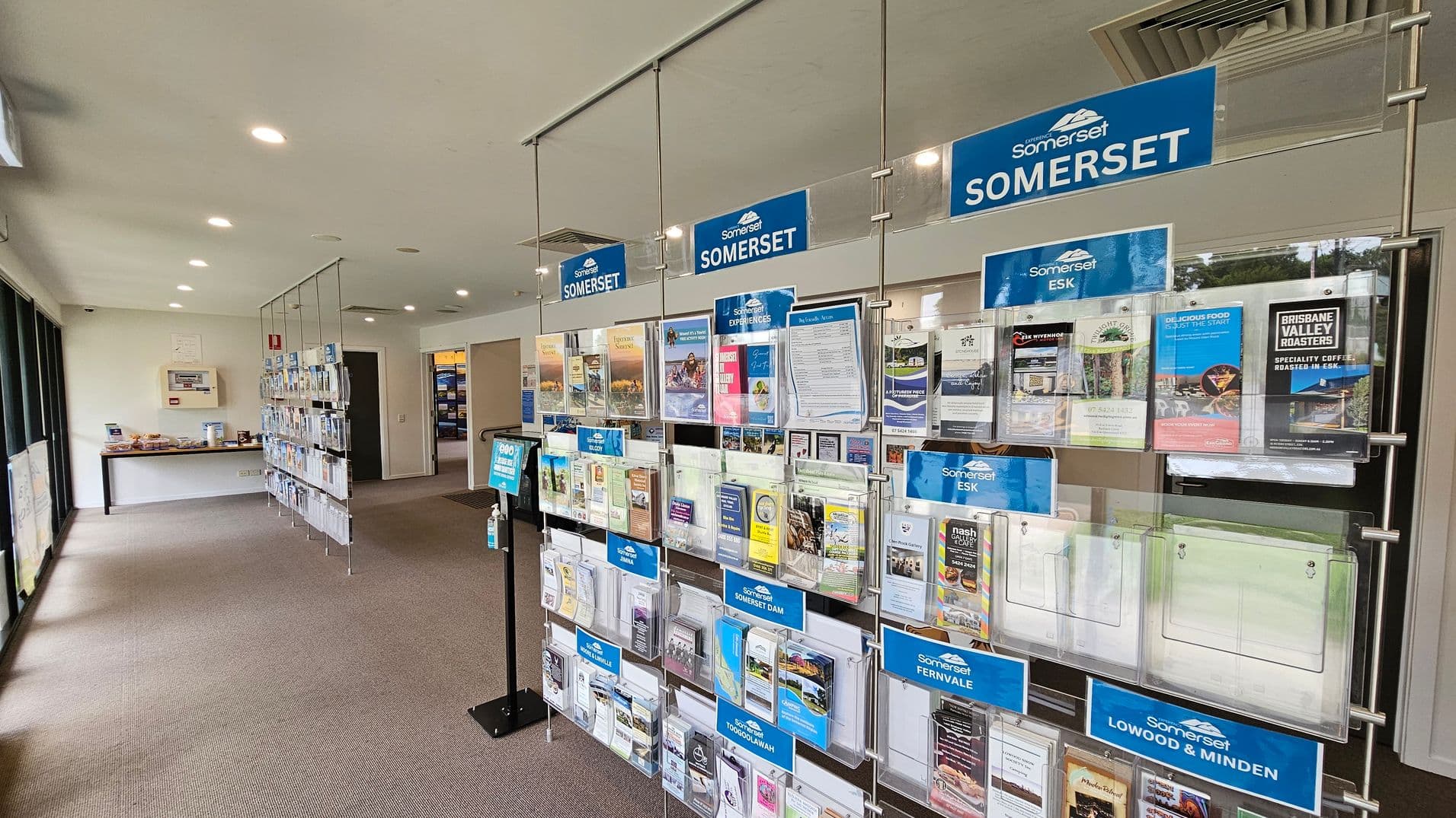 On the left, there are large windows allowing natural light into the room. In the middle, a display rack holds various brochures and pamphlets, with blue signs labelled "Somerset" and other locations like "Esk" and "Fernvale". On the right, more brochures are visible. The floor is carpeted, and the ceiling has recessed lighting. A table with additional materials is in the background.