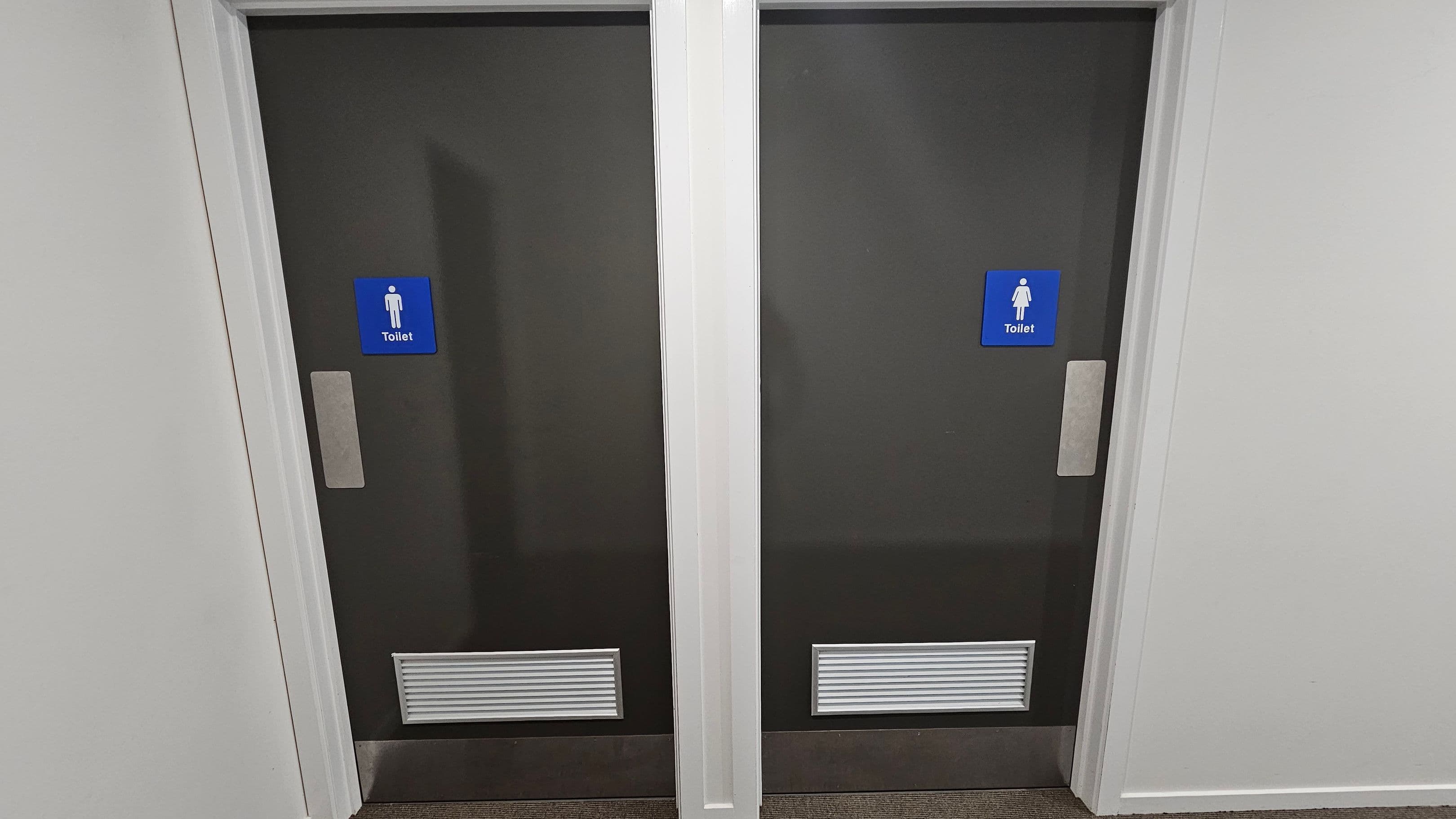 Two grey doors with blue signs indicating toilets. The left door has a male symbol, and the right door has a female symbol. Both doors have silver handles and ventilation grilles at the bottom. The walls surrounding the doors are white, and the floor is covered with a brown carpet.