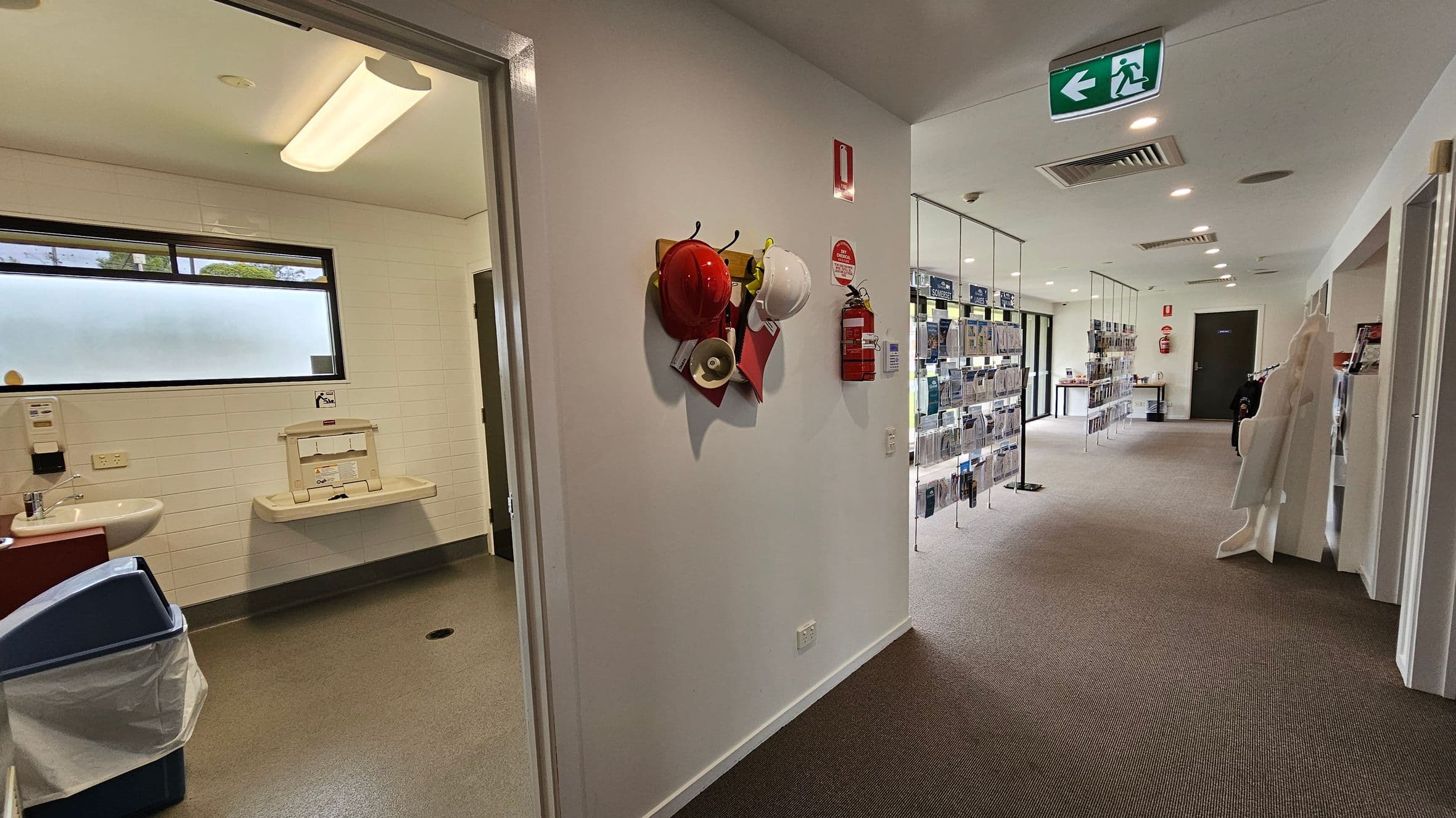 On the left, there's a room with a sink, a baby changing station, and a waste bin. The wall is tiled, and there's a frosted window. In the middle, a wall displays red and white hard hats, a megaphone, and a fire extinguisher. On the right, a corridor with brochures on racks and a brown carpet leads to a door. The ceiling has recessed lights and an exit sign.