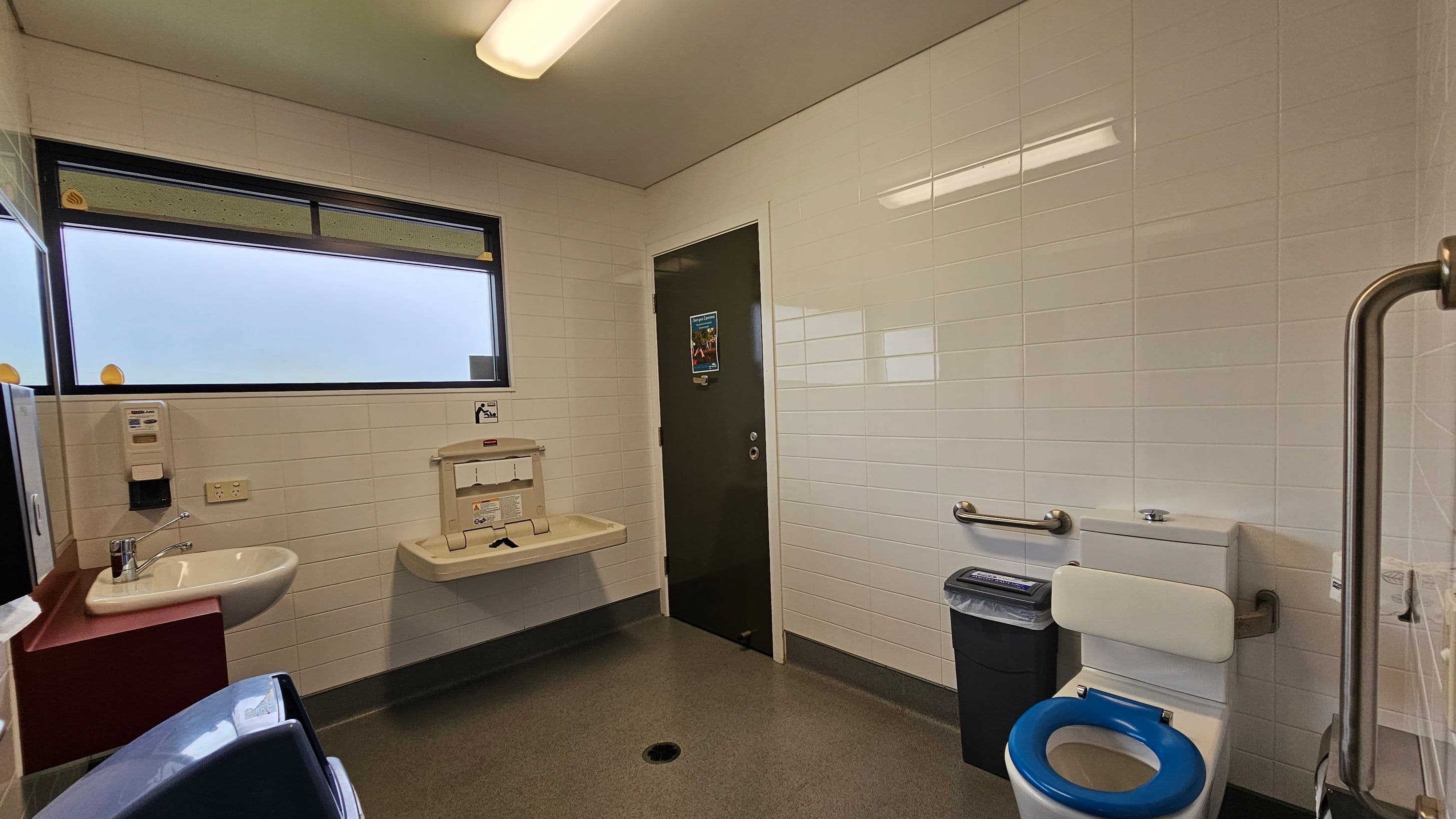 A public restroom with white tiled walls and a grey speckled floor. On the left, there's a sink with a soap dispenser and a hand dryer nearby. Above the sink is a frosted window. In the centre, a baby changing station is mounted on the wall. On the right, a toilet with a blue seat is equipped with support rails, and a waste bin is positioned next to it. A green door with a poster is in the background. The ceiling has a fluorescent light fixture.