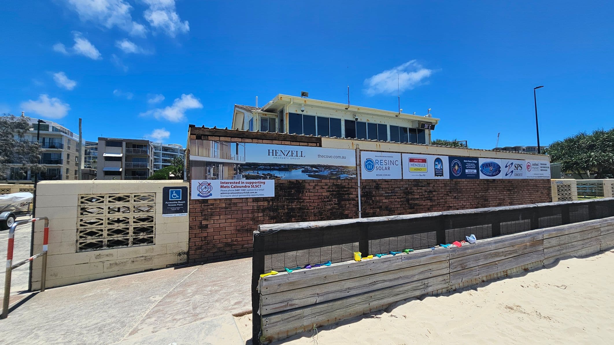 A sandy beach in the foreground with a wooden barrier topped with colourful plastic toys. Behind the barrier is a brick wall with various signs and advertisements, including logos for Henzell Property Group and Resinc Solar. Above the wall is a two-storey building with large windows. To the left, there are modern apartment buildings with balconies.