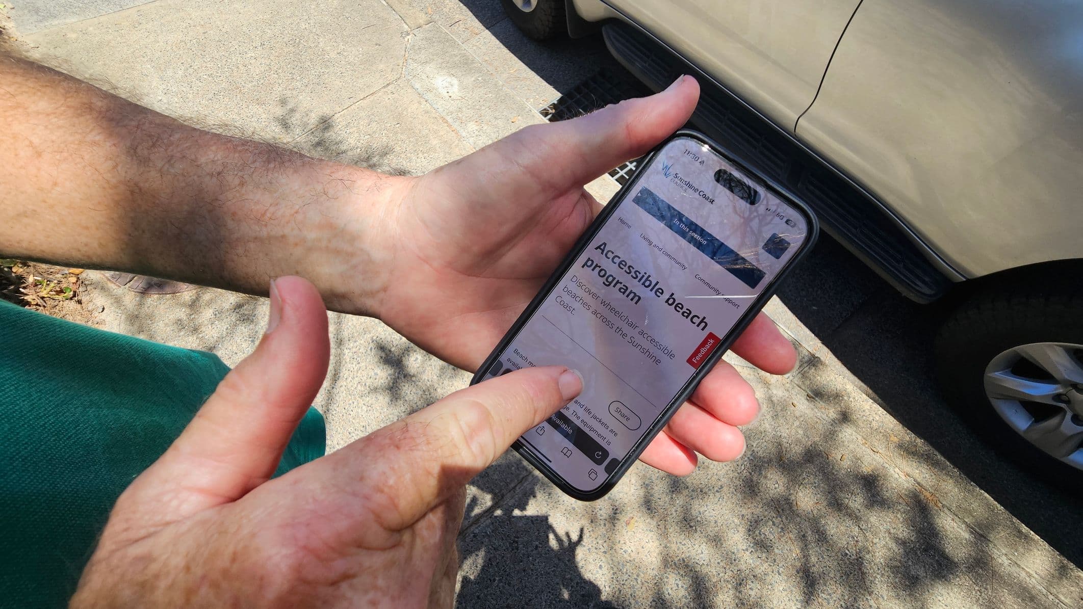 A person is holding a smartphone in their hands, with the screen displaying information about an "Accessible beach program." The person's left hand is supporting the phone, while the right hand is pointing at the screen. In the background, part of a light-coloured vehicle is visible, parked on a concrete surface. Shadows of leaves are cast on the ground, suggesting nearby trees.