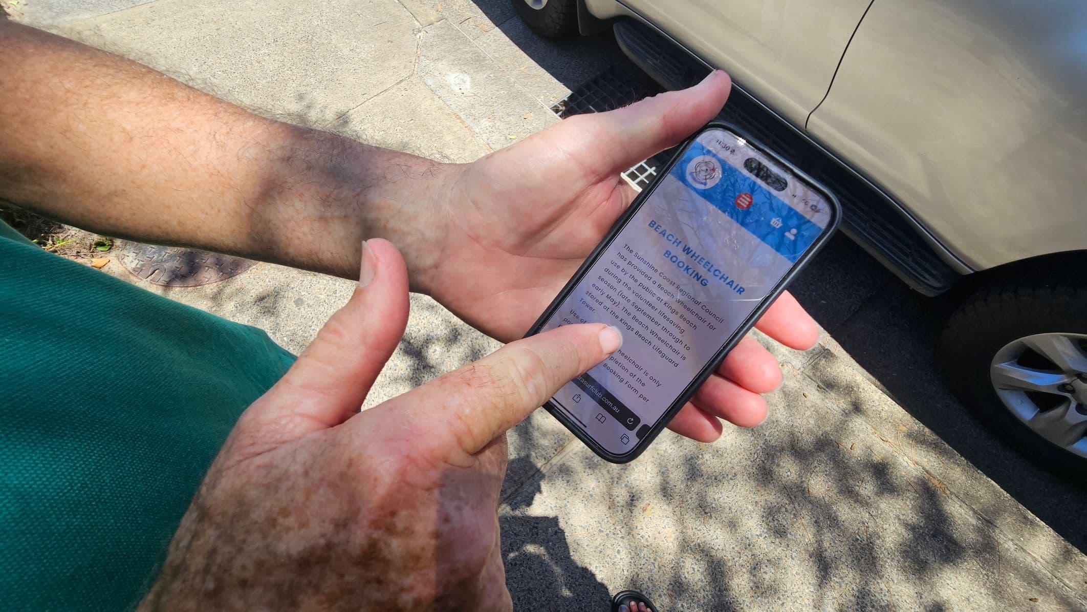 A person is holding a smartphone in their left hand, with their right hand pointing at the screen. The phone displays a webpage titled "Beach Wheelchair Booking." The person is wearing a green shirt. In the background, there is a light-coloured car parked on a paved surface, with shadows of tree branches visible on the ground.