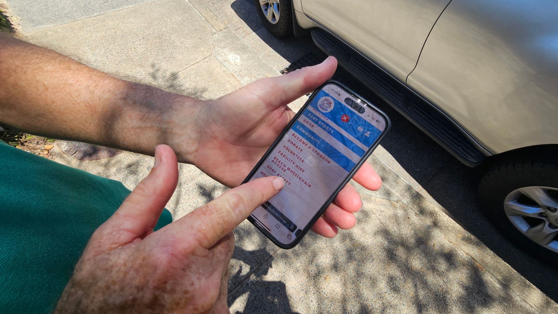 A person holding a smartphone in their left hand, with their right hand pointing at the screen. The phone displays a website with options like "Become a Sponsor" and "Volunteer". The background shows a pavement and part of a parked silver car. Shadows of leaves are visible on the ground.