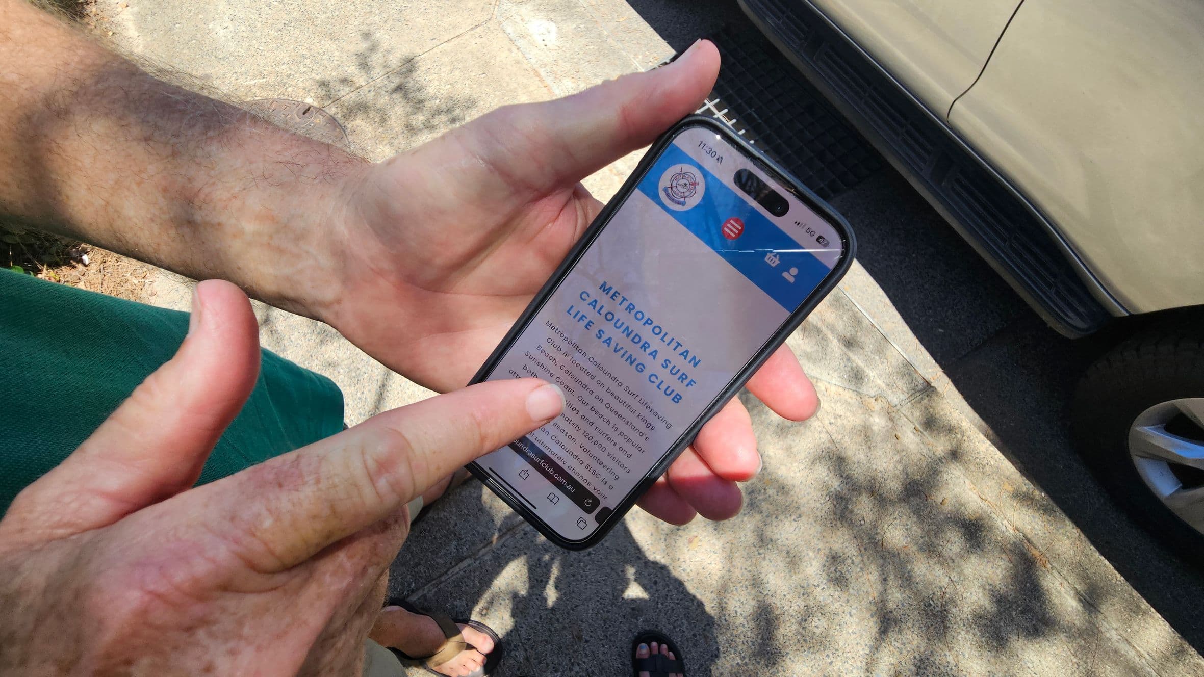 A person is holding a smartphone displaying a webpage for the Metropolitan Caloundra Surf Life Saving Club. The phone is held in one hand, while the other hand points at the screen. The background shows a concrete pavement with shadows from nearby trees. Part of a parked car is visible on the right side of the image. The person is wearing a green shirt, and their feet, in sandals, are partially visible at the bottom.
