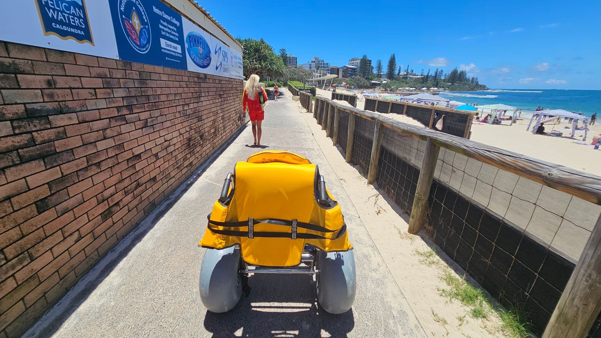A pathway with a brick wall on the left displaying various signs. A person in a red outfit walks ahead. In the centre, a yellow beach wheelchair with large grey wheels is visible. On the right, a wooden fence separates the path from a sandy beach with people, umbrellas, and the ocean in the background.
