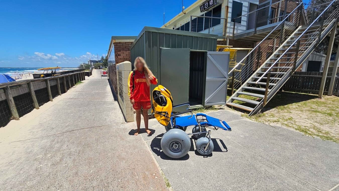 A person in a red lifeguard uniform stands on a concrete path next to a yellow and blue beach wheelchair. To the right, there's a green metal shed with open doors and a staircase leading up to a building. On the left, a wooden fence separates the path from the sandy beach, where a yellow lifeguard vehicle and beachgoers are visible.