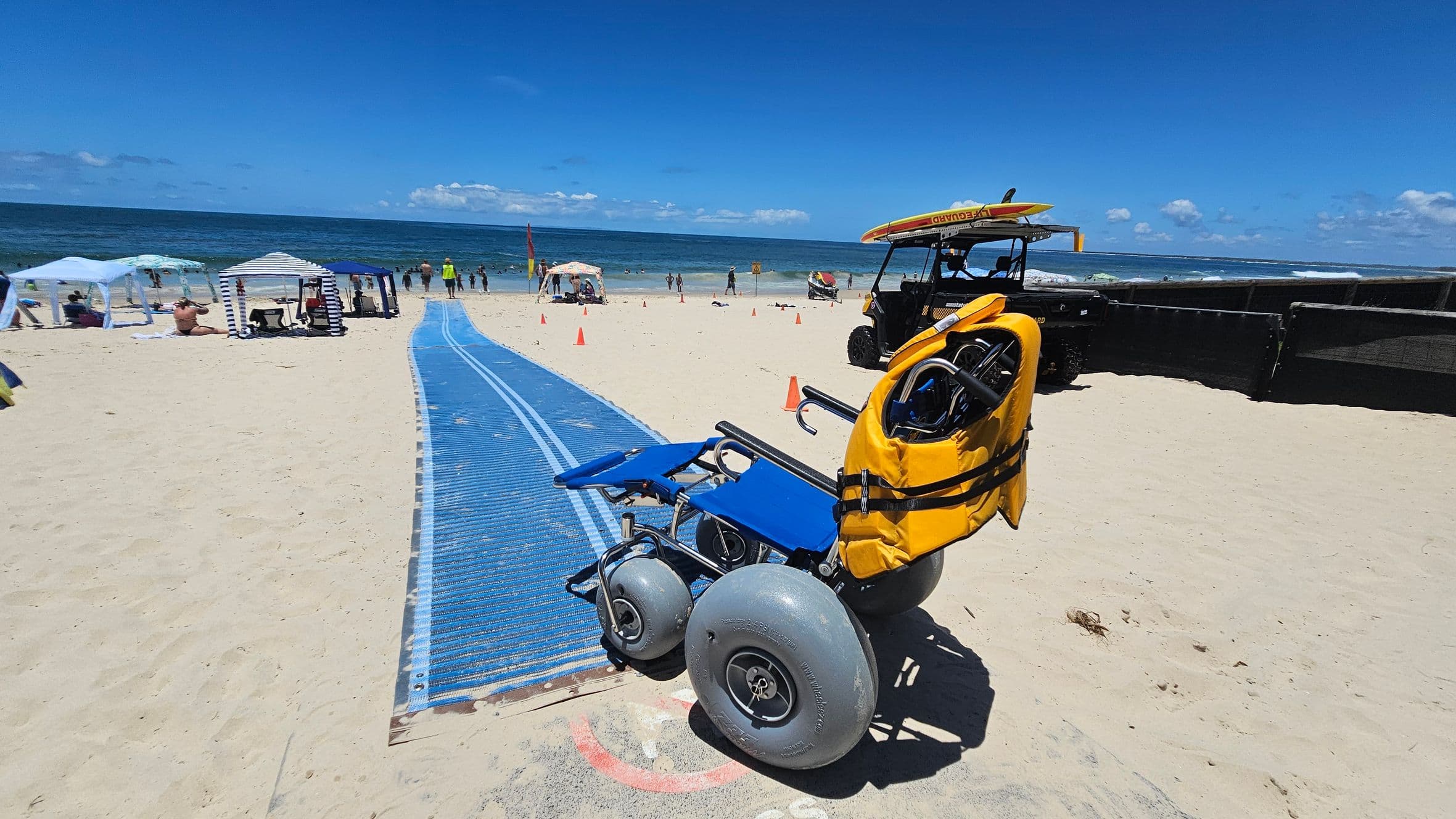 A sandy beach with a blue mat pathway leading towards the sea. On the right, a beach wheelchair with large grey wheels and a yellow seat is parked. In the background, people are under white and striped canopies near the water. A lifeguard vehicle with a surfboard on top is parked further back. Orange cones are scattered around the area. The ocean is visible in the distance with small waves.