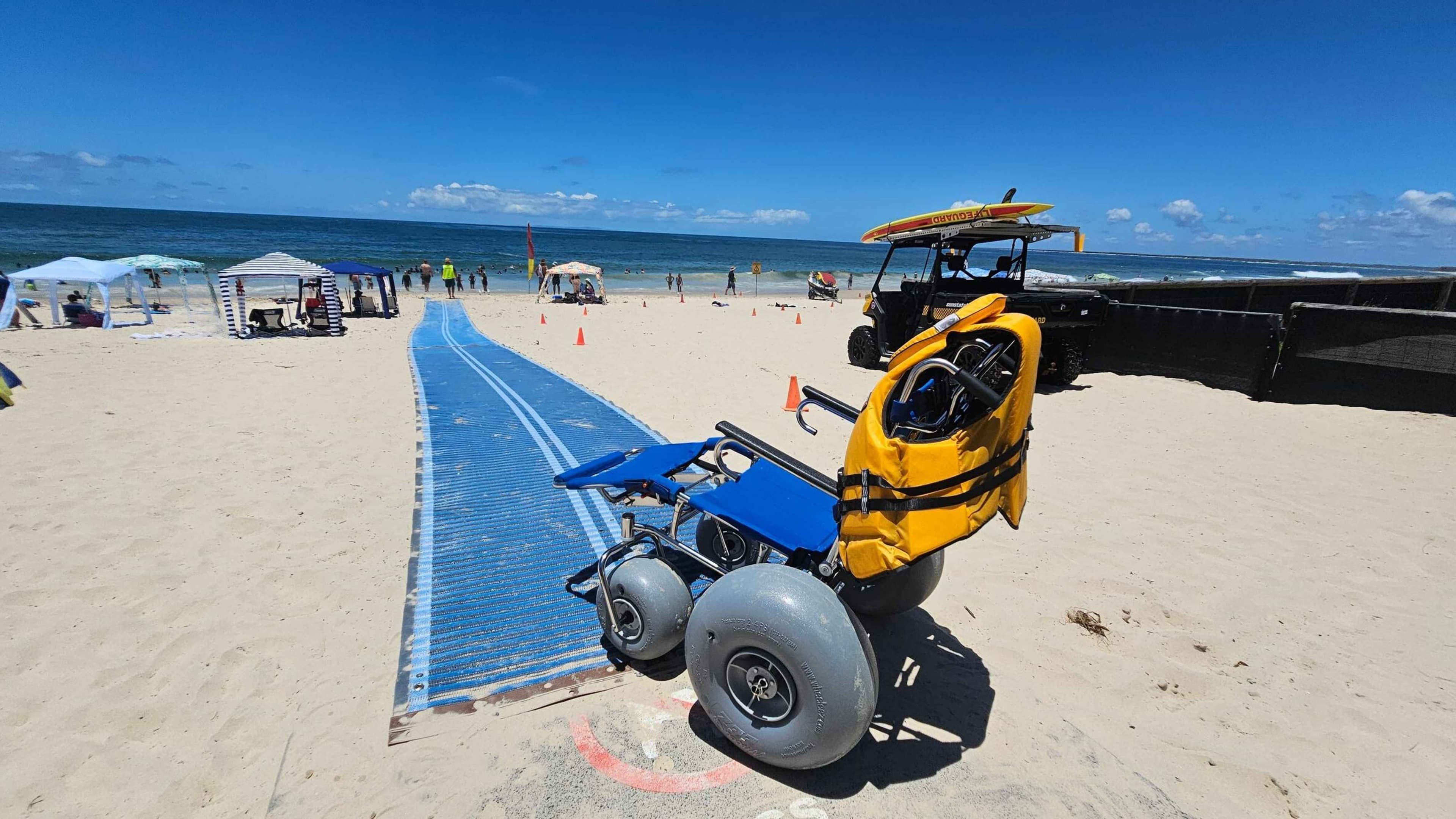 A sandy beach with a blue mat pathway leading towards the ocean. On the left, a blue and yellow tent is set up near several beachgoers under umbrellas. On the right, a lifeguard vehicle with a surfboard on top is parked on the sand. Orange cones are placed along the pathway. The ocean is in the background with people swimming and enjoying the water.