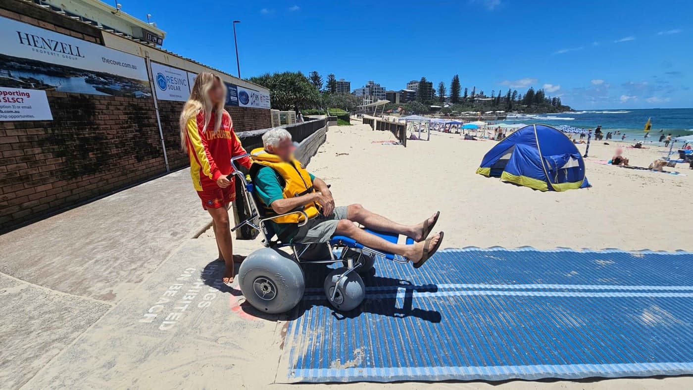 A lifeguard in a red and yellow uniform stands on the left, pushing a man in a beach wheelchair with large wheels. The man is wearing a yellow life jacket and sitting comfortably. They are on a sandy beach with a blue mat leading towards the sea. In the background, there is a blue tent and several people enjoying the beach. A brick wall with advertisements is on the left side. The ocean is visible on the right, with waves and people swimming.