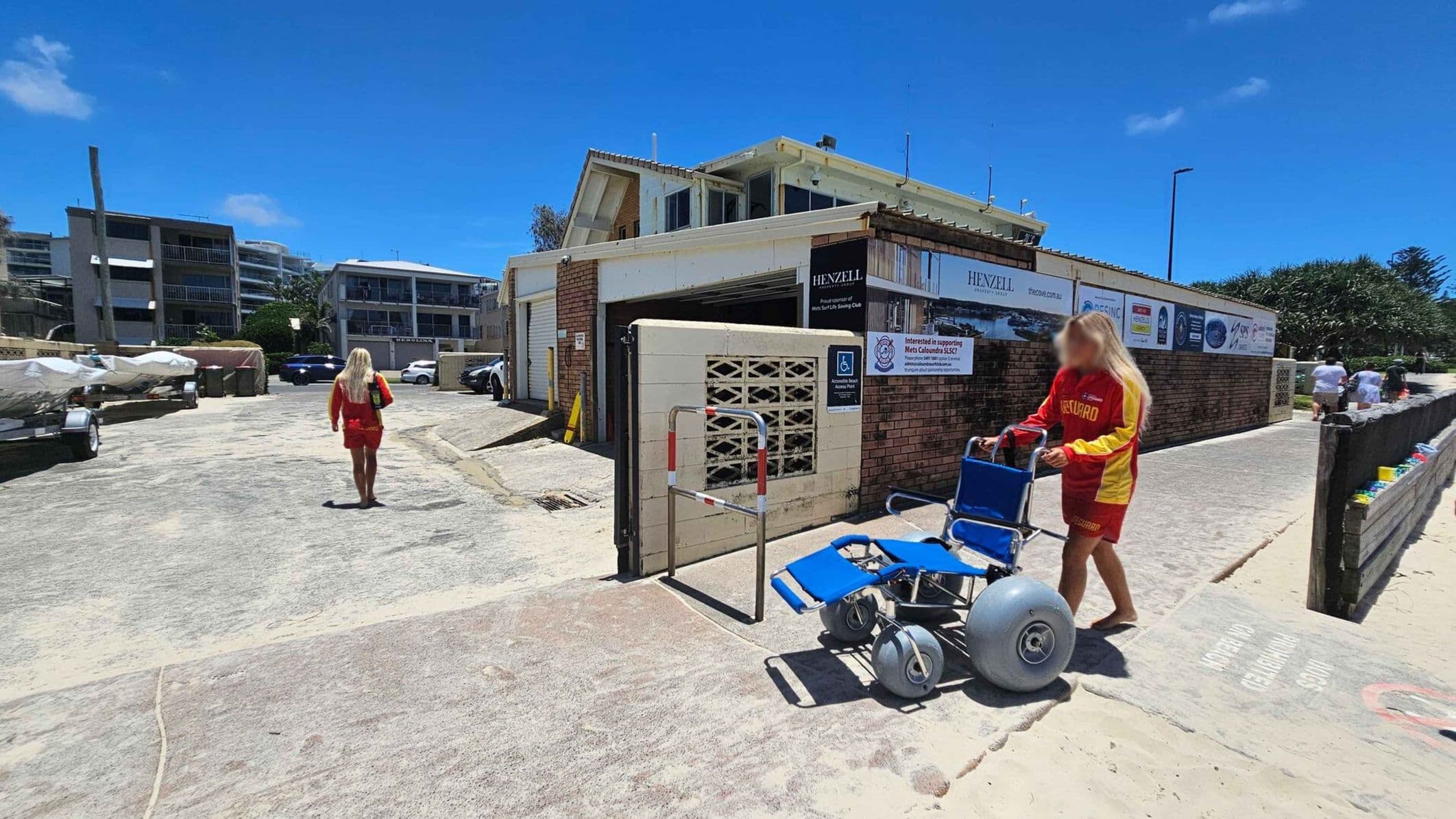 A lifeguard in a red and yellow uniform is pushing a blue beach wheelchair with large wheels on the right. Another lifeguard is walking away in the background on the left. The scene is set near a building with signs and advertisements, and there are several parked cars and boats on trailers. The ground is sandy, and a wooden fence lines the path on the right.
