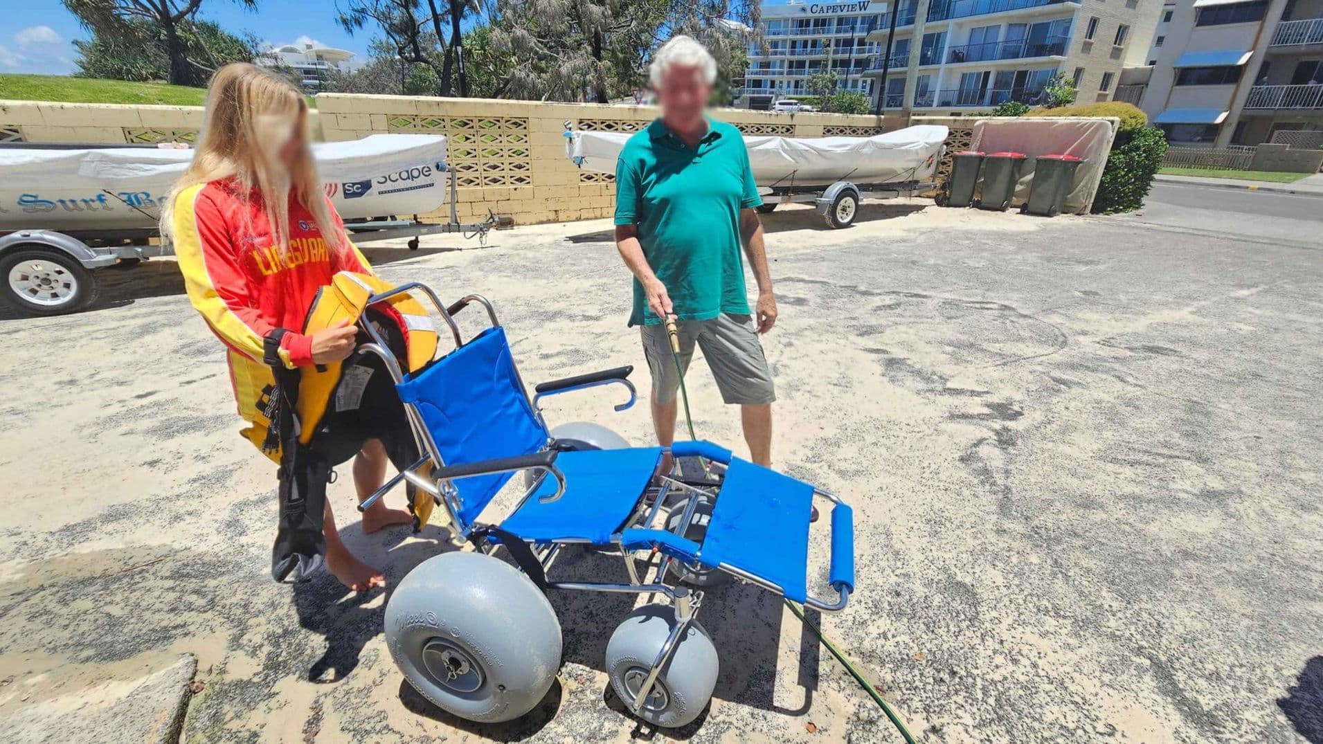 A woman in a red and yellow lifeguard uniform stands on the left, holding a life jacket. A man in a green shirt and grey shorts is on the right, holding a blue beach wheelchair with large wheels. In the background, there are boats on trailers, a brick wall, and a building with balconies. The ground is sandy with patches of green.