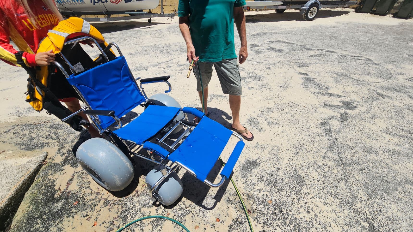 A blue beach wheelchair with large grey wheels is on a sandy surface. On the left, a person in a red and yellow lifeguard uniform holds the chair. On the right, a person in a green shirt and grey shorts stands, holding a hose. The background shows a trailer and some equipment. The ground is a mix of sand and patches of darker material.