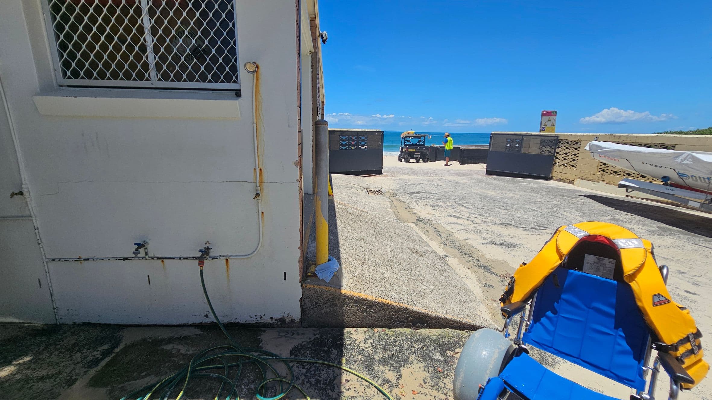 A building with a barred window and two water taps is on the left. A green hose lies on the ground. In the centre, a person in a bright shirt stands near a utility vehicle by the sea. On the right, a blue and yellow beach wheelchair is visible, and a covered boat rests on a trailer. The ground is a mix of concrete and sand, and a low wall separates the area from the beach.